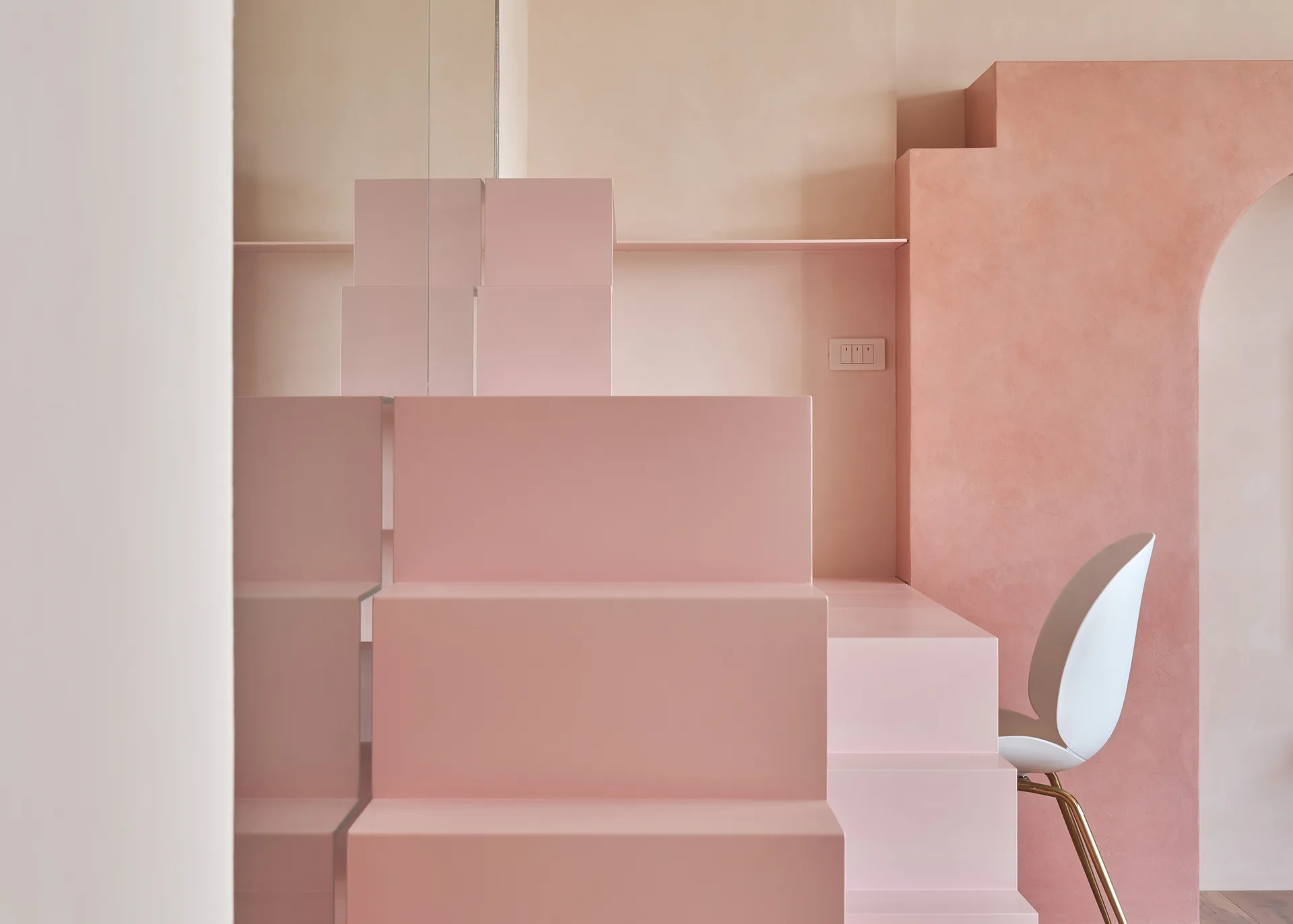 A minimalist pink stepped platform sits next to a textured salmon-colored plaster archway and a modern white chair.