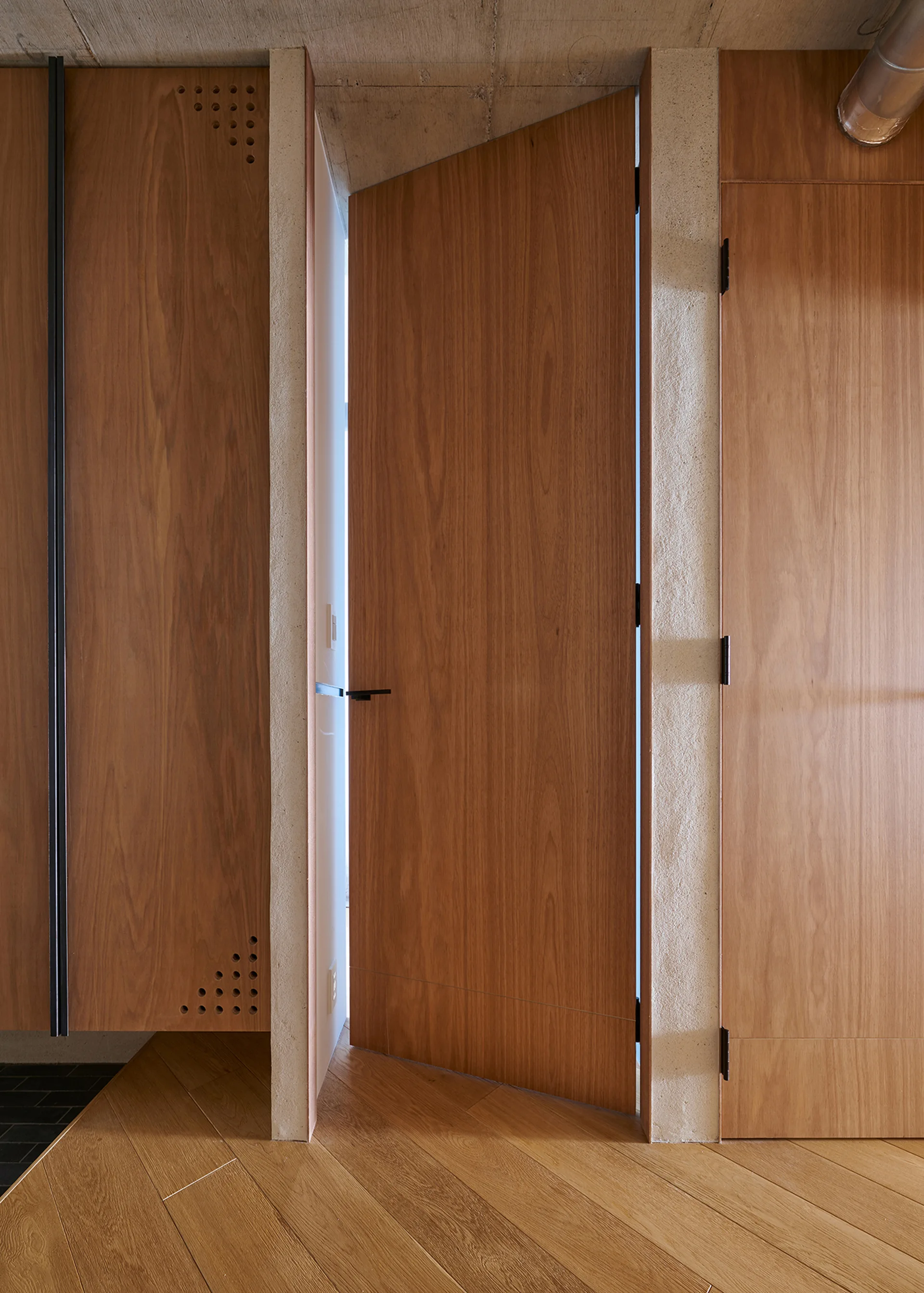 A custom angled wood door stands slightly open between vertical concrete wall pillars under an exposed concrete ceiling.