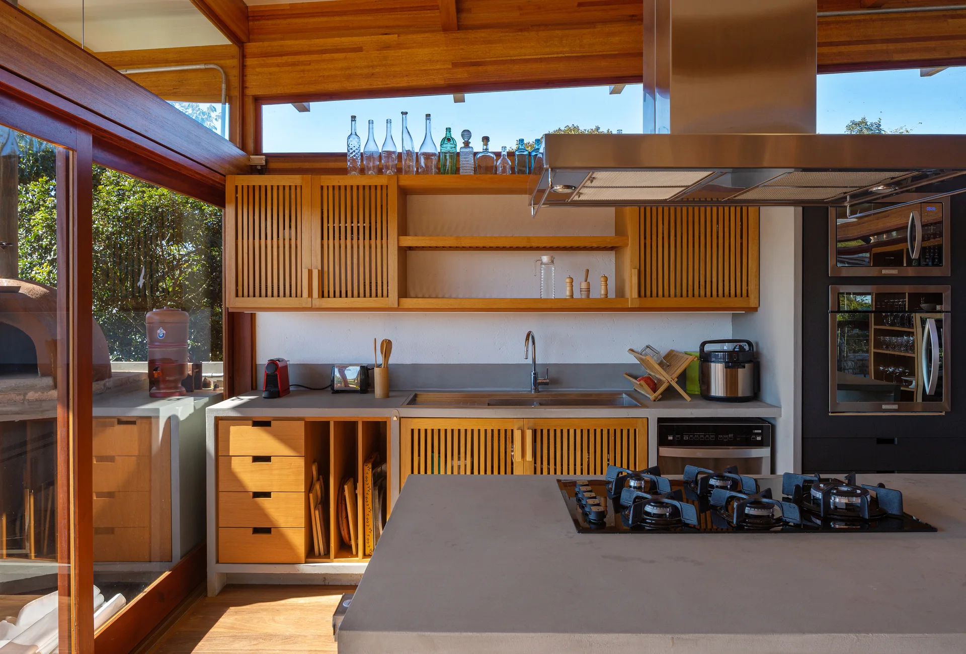 A modern kitchen features a concrete island with a gas cooktop, slatted wood cabinetry, a stainless steel sink, and a clerestory window.