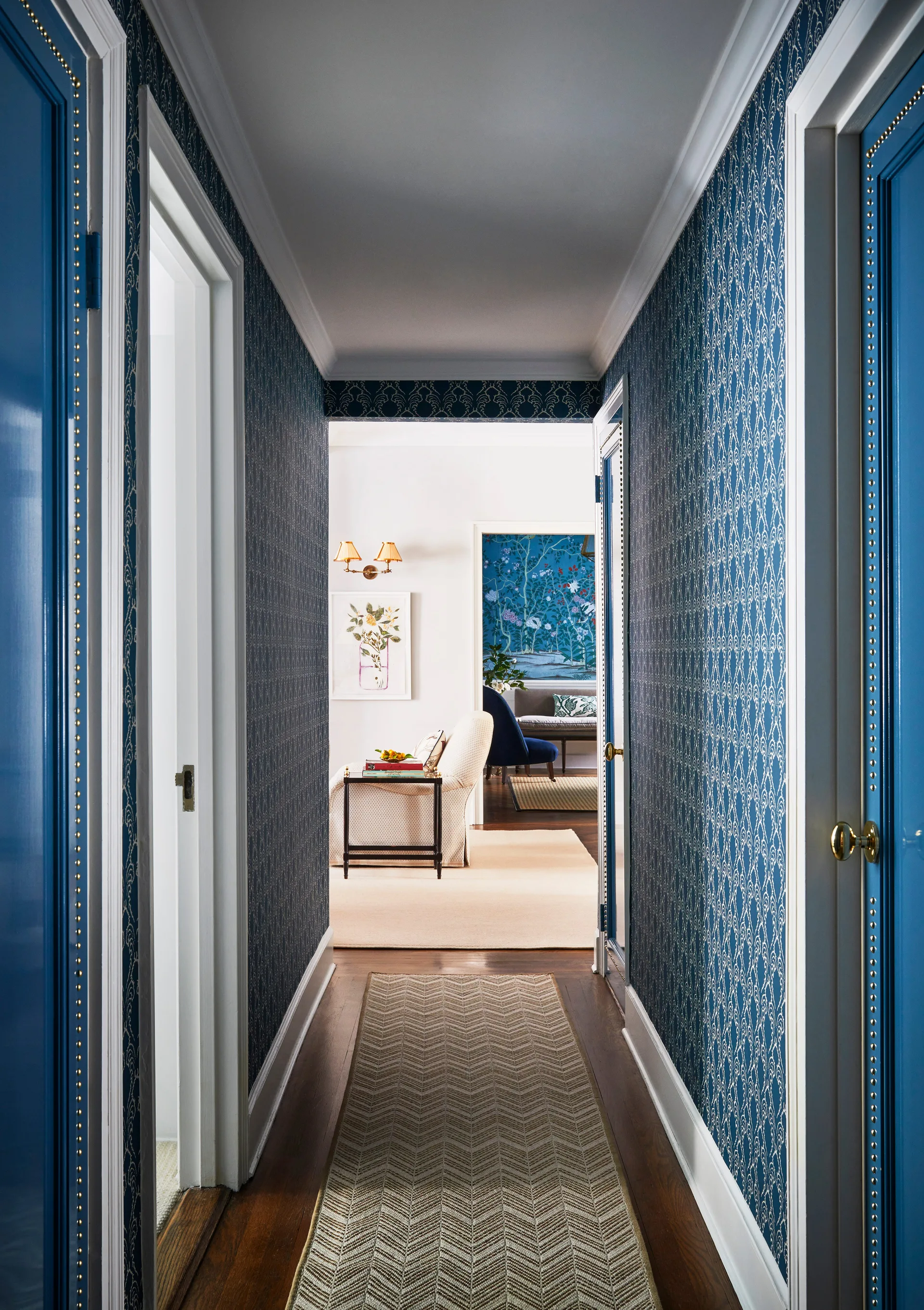 A narrow hallway featuring blue patterned wallpaper, high-gloss blue doors with gold nailhead trim, and a patterned runner rug over dark wood floors.