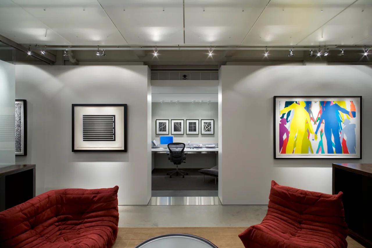 A modern office lounge features two red ribbed chairs in the foreground, white walls with framed art, and an opening to a back office workspace.