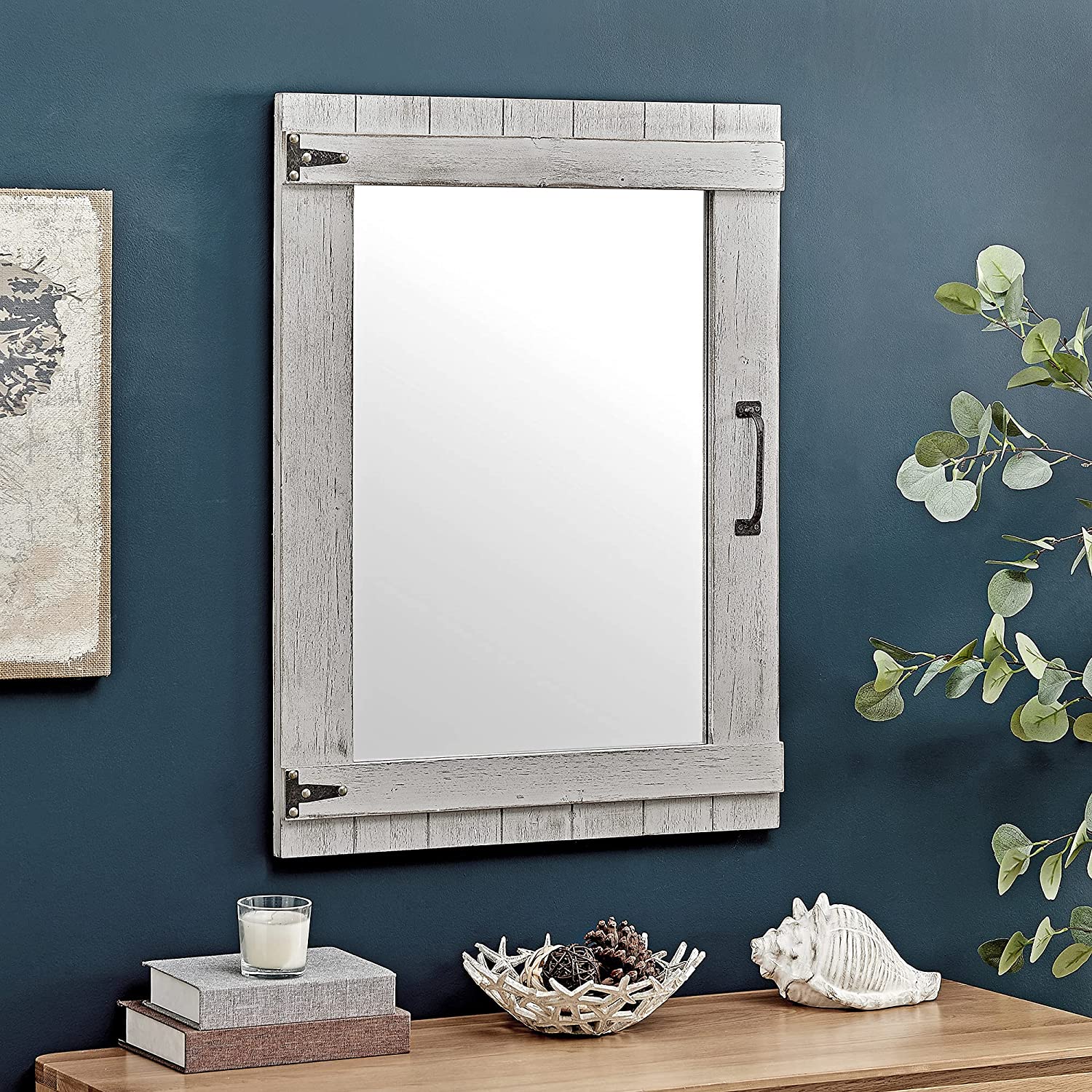 A rectangular whitewashed wood mirror with black iron hinges hangs on a deep teal wall above a tabletop displaying coastal decor.