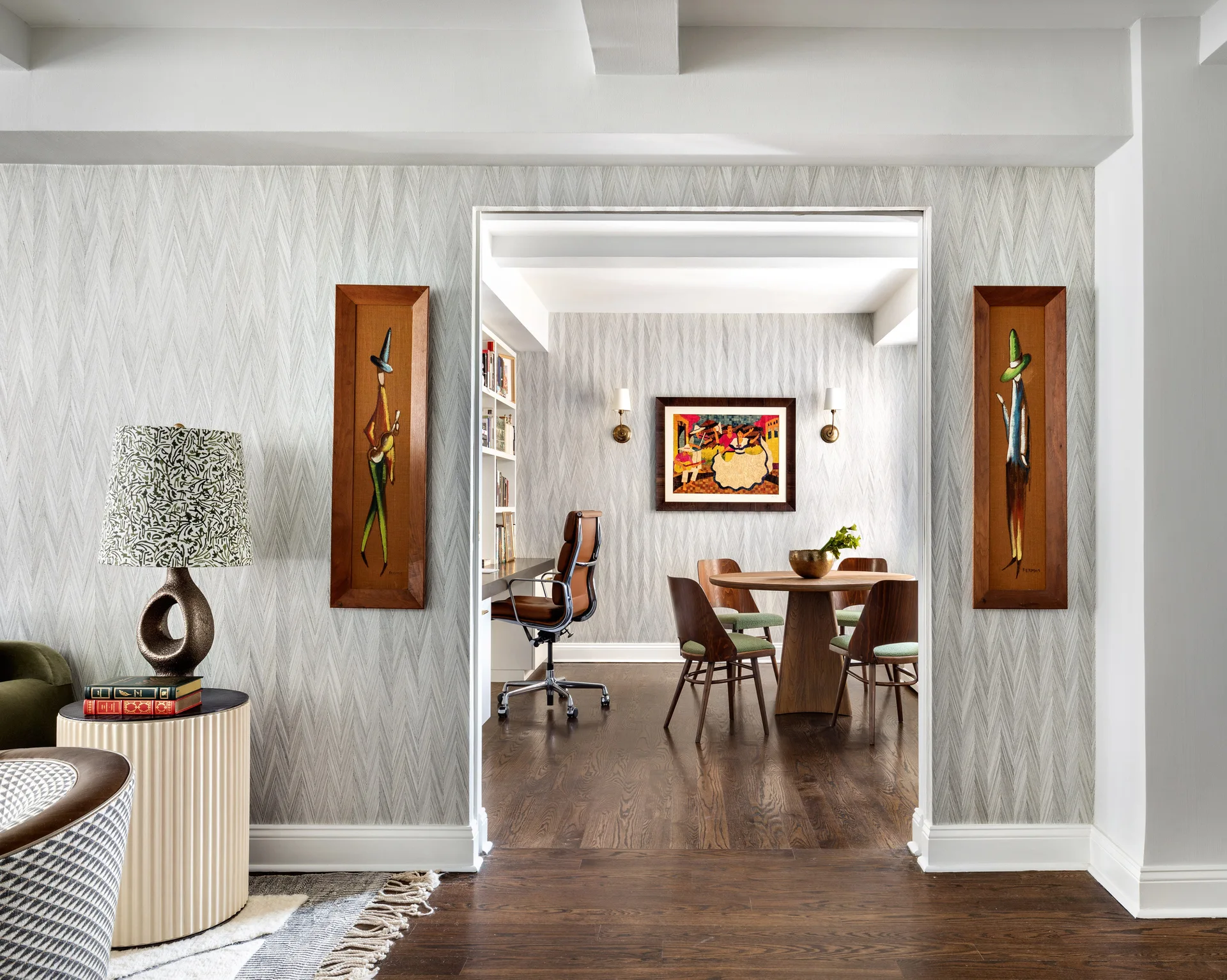 A view through a wide doorway reveals a dining room and home office with chevron wallpaper, a round wooden table, and mid-century chairs.