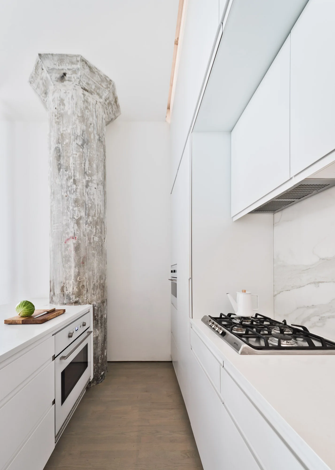 A minimalist white kitchen featuring flat-front cabinets, a gas stovetop, and a prominent exposed concrete column next to a light wood floor.
