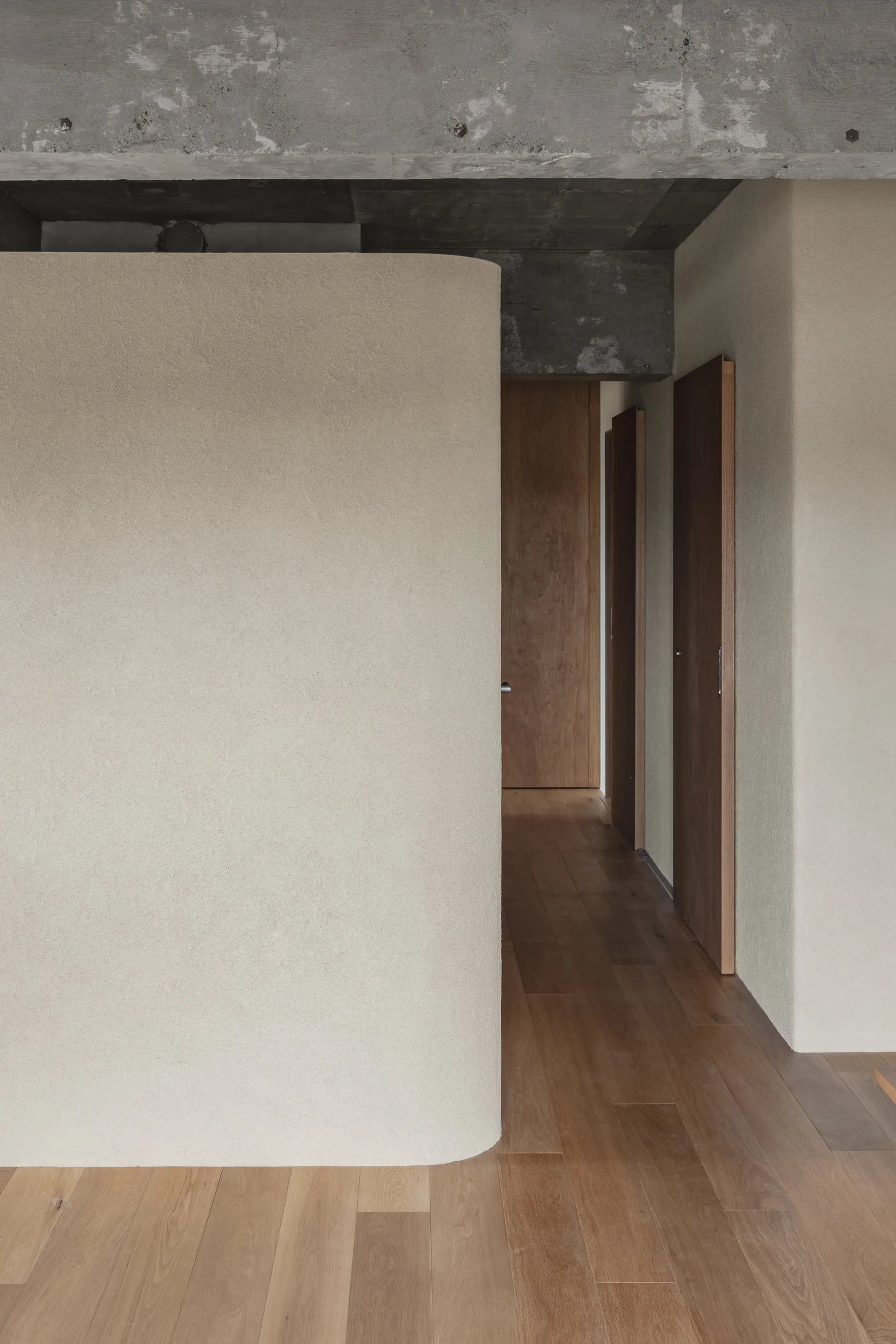 A minimalist hallway features a curved beige plaster wall, natural wood flooring and doors, and an exposed concrete ceiling beam.