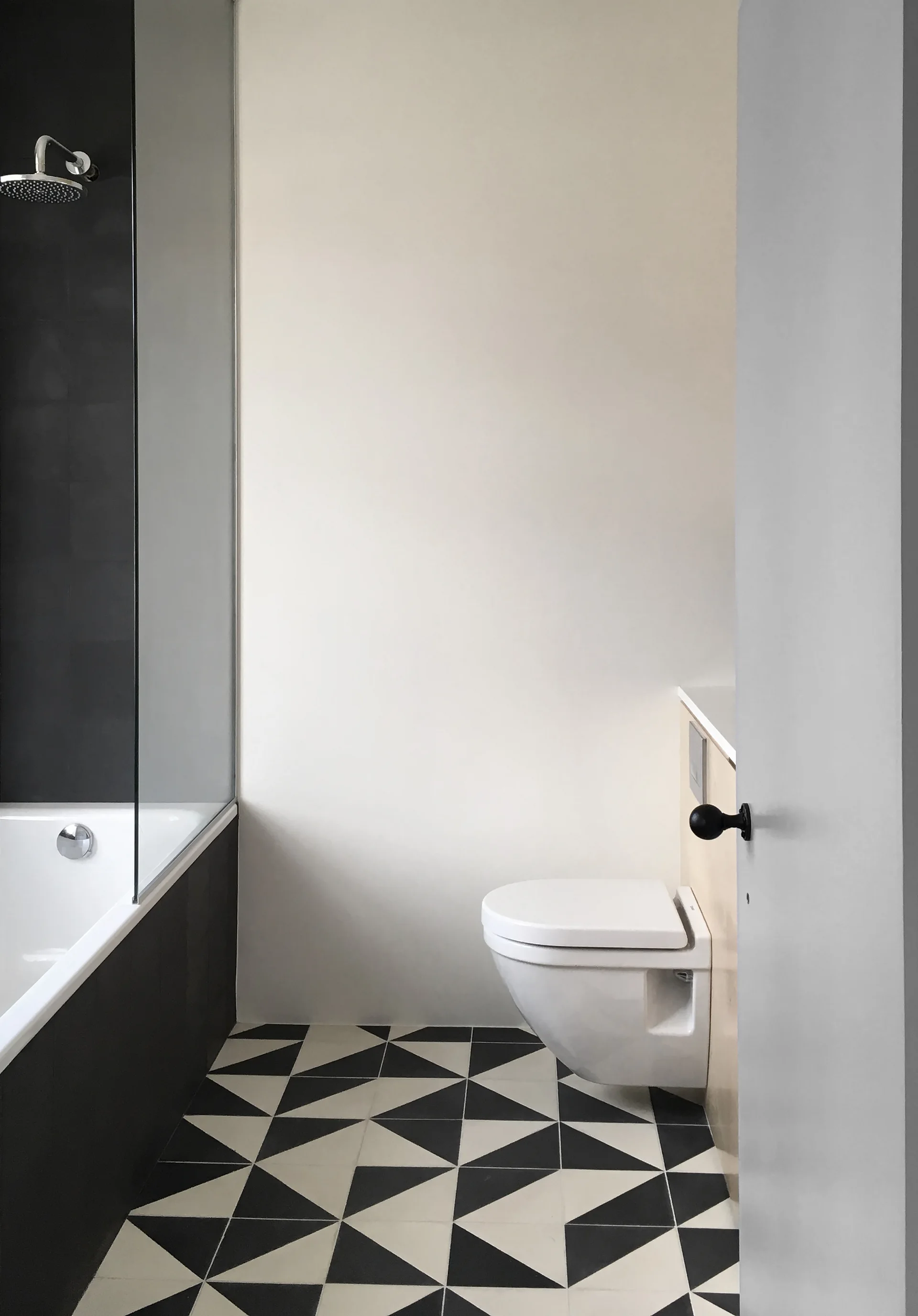 A modern bathroom features a white bathtub, a wall-mounted toilet with a light wood casing, and high-contrast black and white geometric floor tiles.