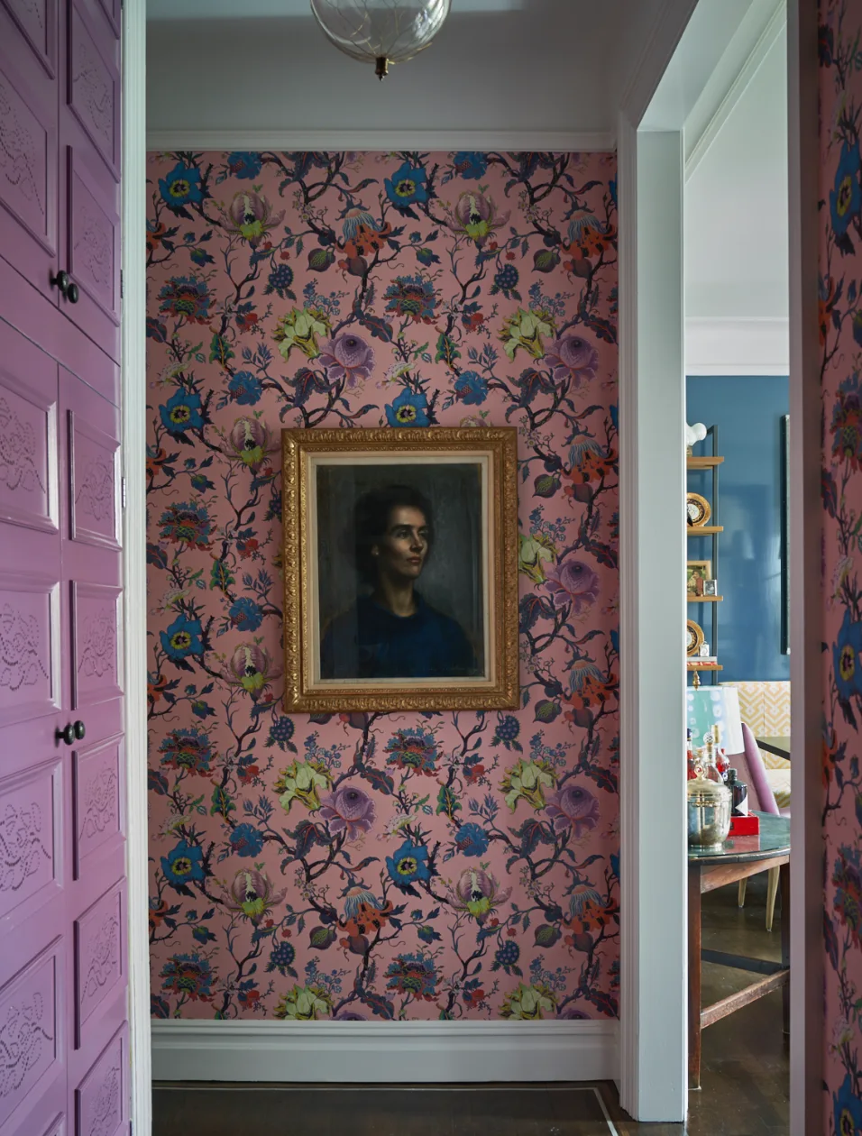 A narrow hallway features vibrant pink floral wallpaper, a gold-framed portrait, and built-in purple cabinets, leading to a blue room beyond.