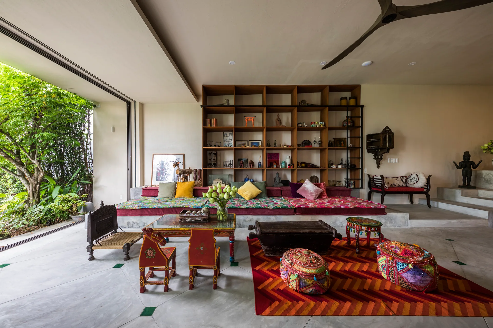 A bohemian living room features a large wooden grid shelving unit, low concrete platform daybeds with colorful textiles, and a red geometric rug.