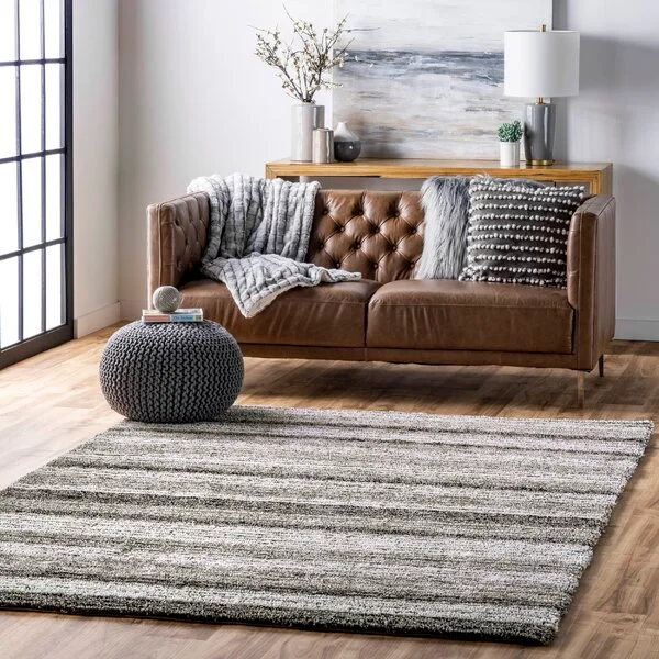 A brown tufted leather sofa sits on a textured, striped grey and white area rug next to a dark grey knitted floor pouf in a bright living room.