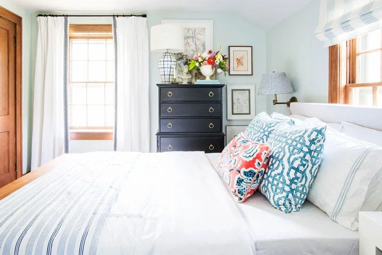 A light blue bedroom featuring a bed with patterned blue and coral pillows next to a black tall chest of drawers with gold handles.