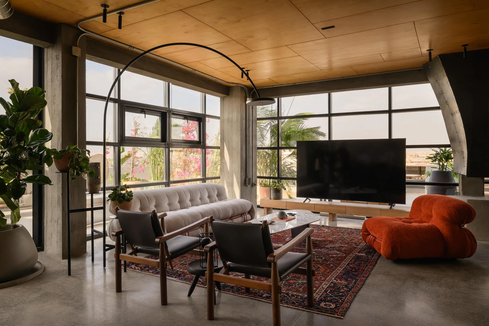 A spacious industrial living room features a tufted sofa, two wooden armchairs, an orange lounge chair, and a large arched floor lamp.