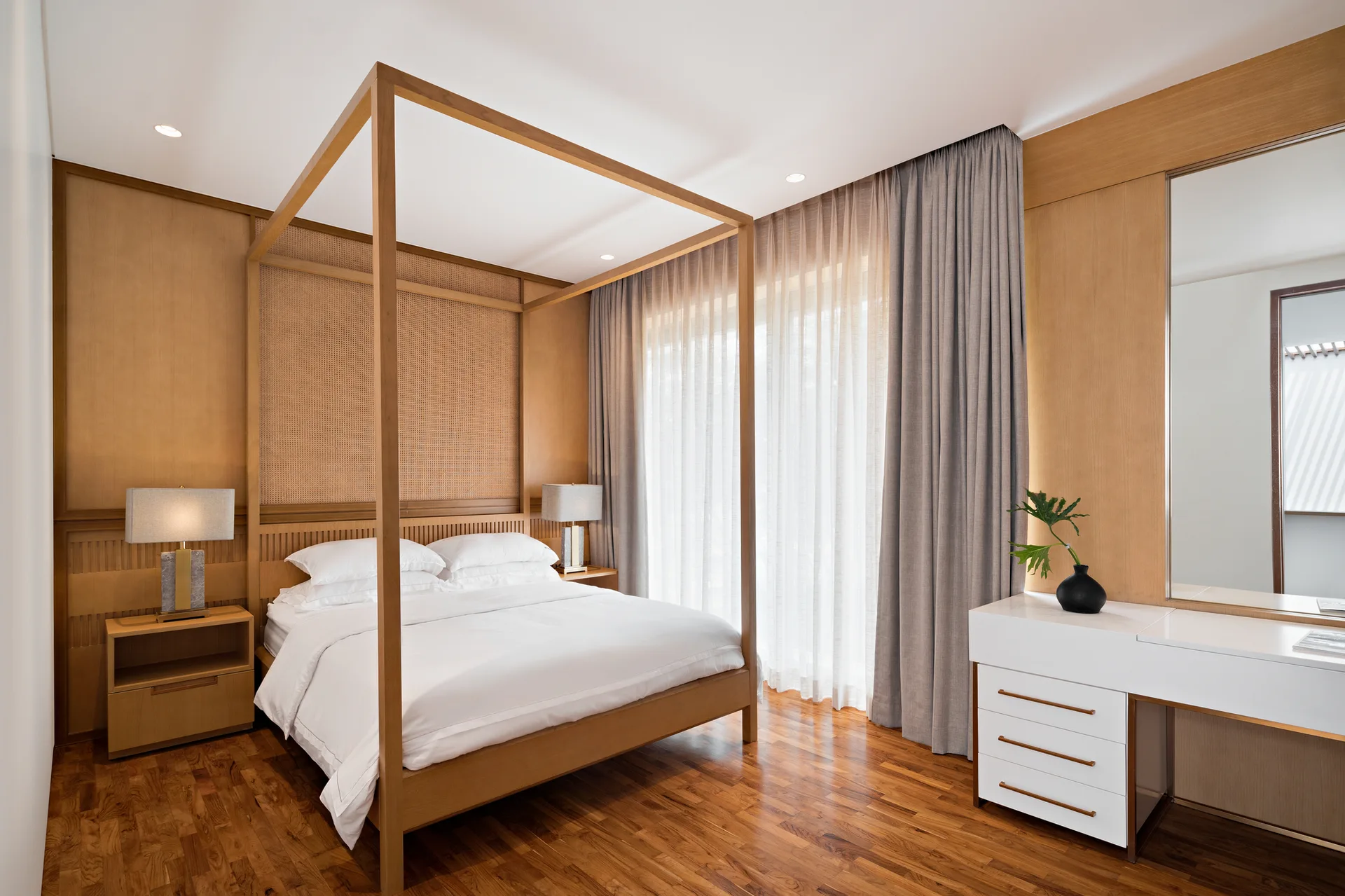 A modern bedroom with a wooden canopy bed, a woven cane headboard, wood-paneled walls, grey curtains, and a white vanity with a large mirror.
