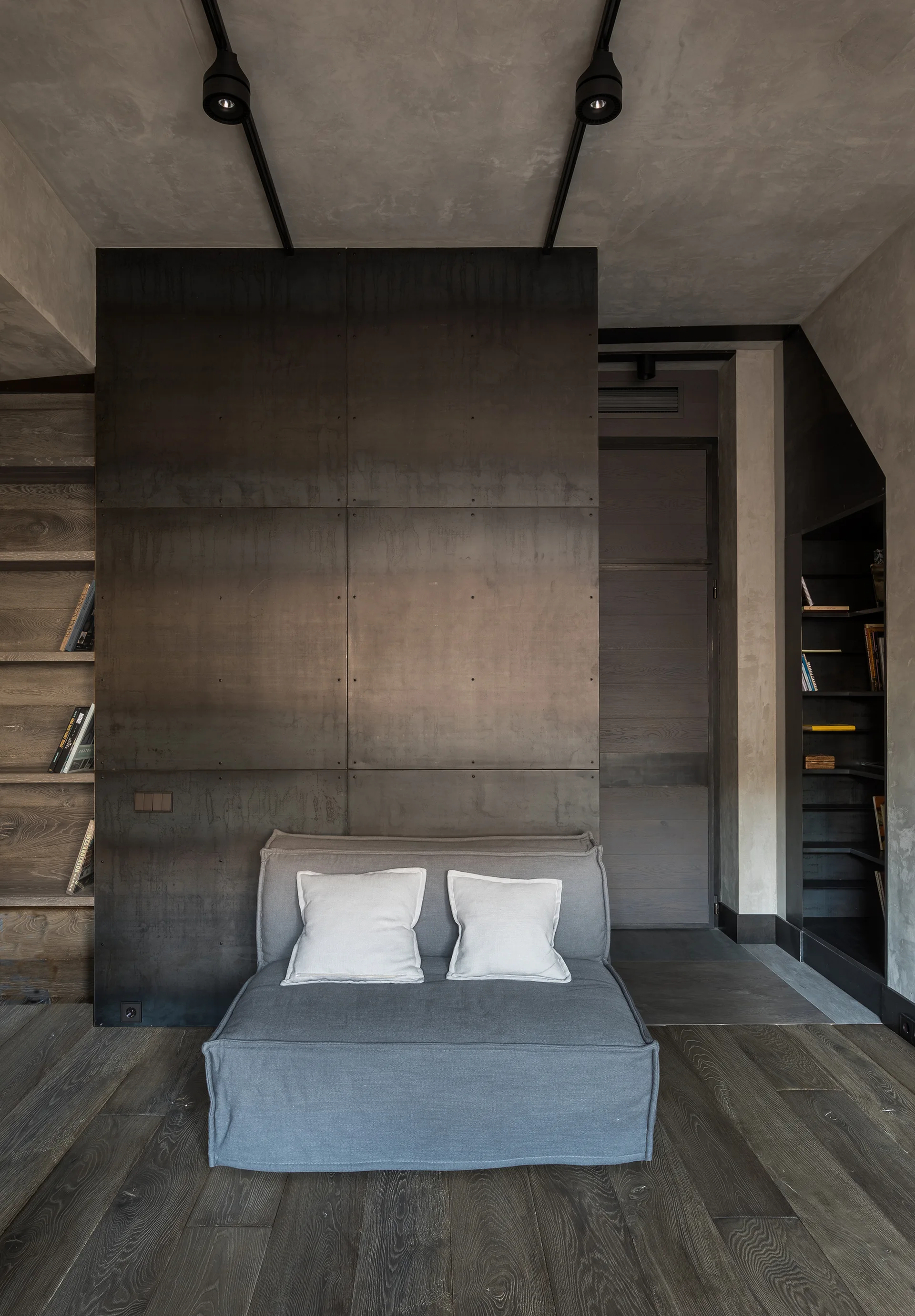 A gray armless sofa with two white cushions sits against a large wall of dark metal panels, flanked by wooden shelves.
