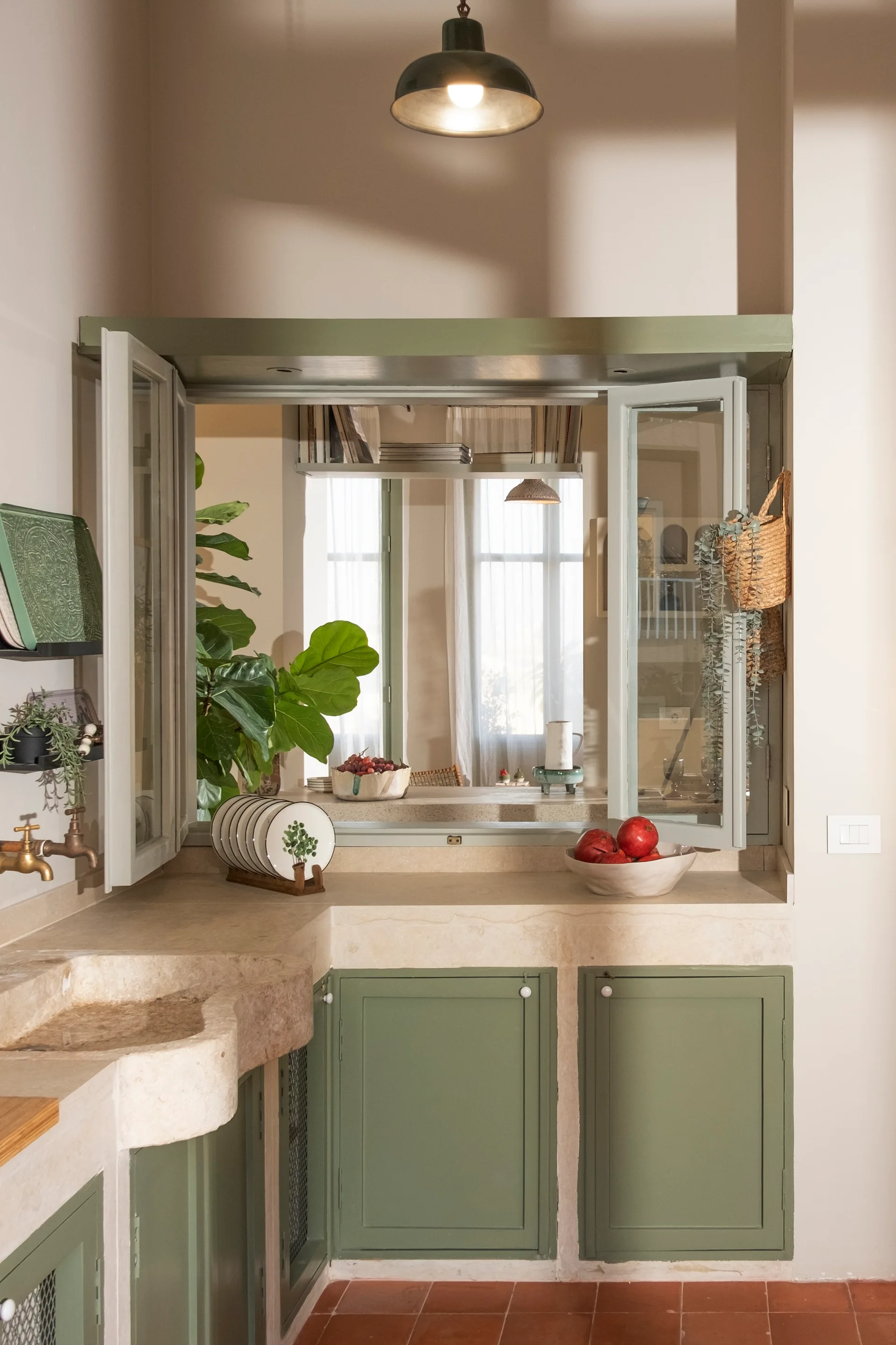A rustic kitchen features sage green cabinetry, a curved stone sink with a brass faucet, terracotta floor tiles, and an interior pass-through window.