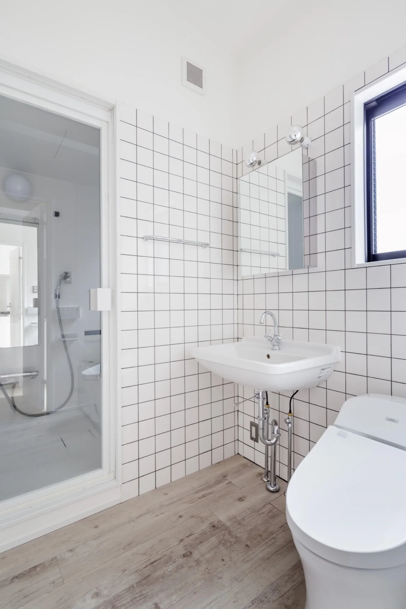 A minimalist bathroom features white square grid tiles, a wall-mounted ceramic sink with exposed chrome pipes, a mirror, and light wood flooring.