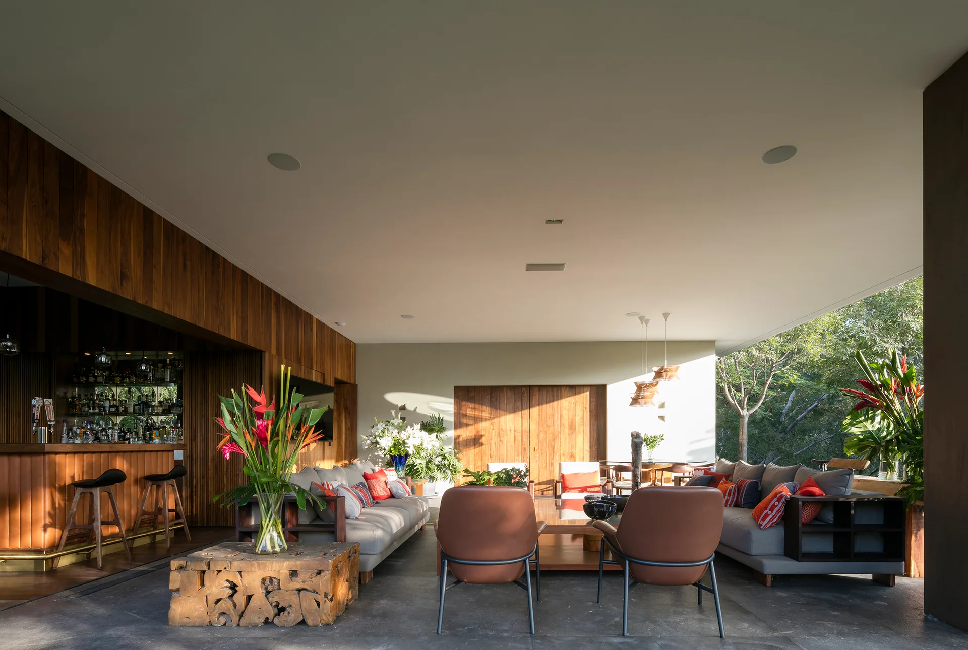 An open-air patio features two grey sofas, brown leather lounge chairs, a wood-paneled bar with stools, and tropical flower arrangements.