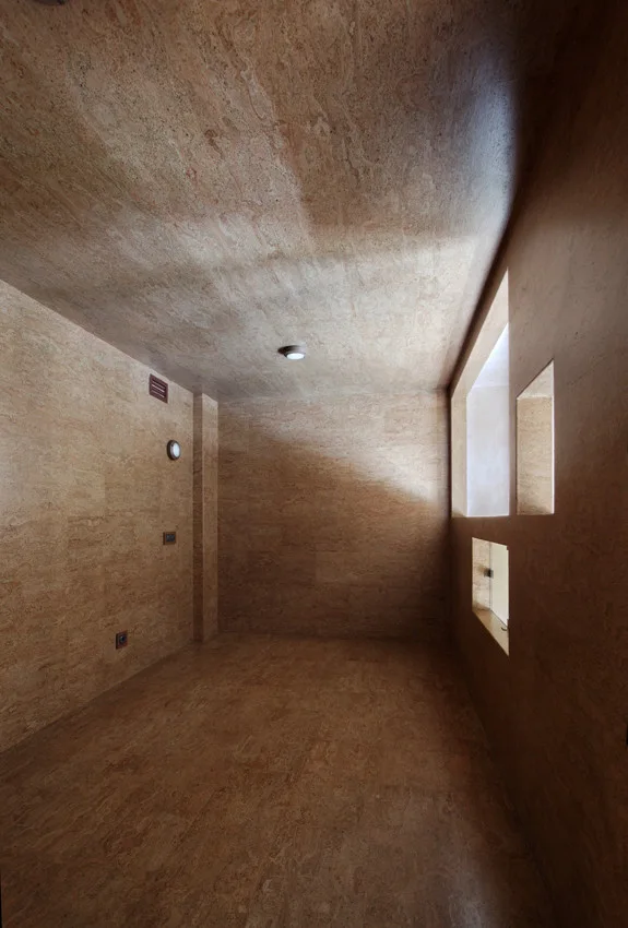 An empty minimalist room with brown textured stone cladding on the floor, walls, and ceiling, featuring small recessed square windows.