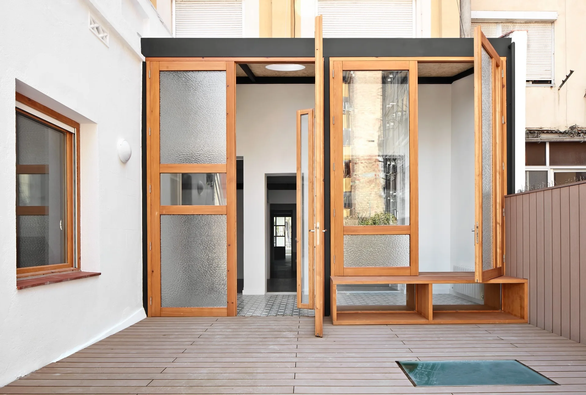 A modern outdoor patio features wood-toned decking, white plaster walls, and large wooden-framed folding glass doors with an integrated bench.