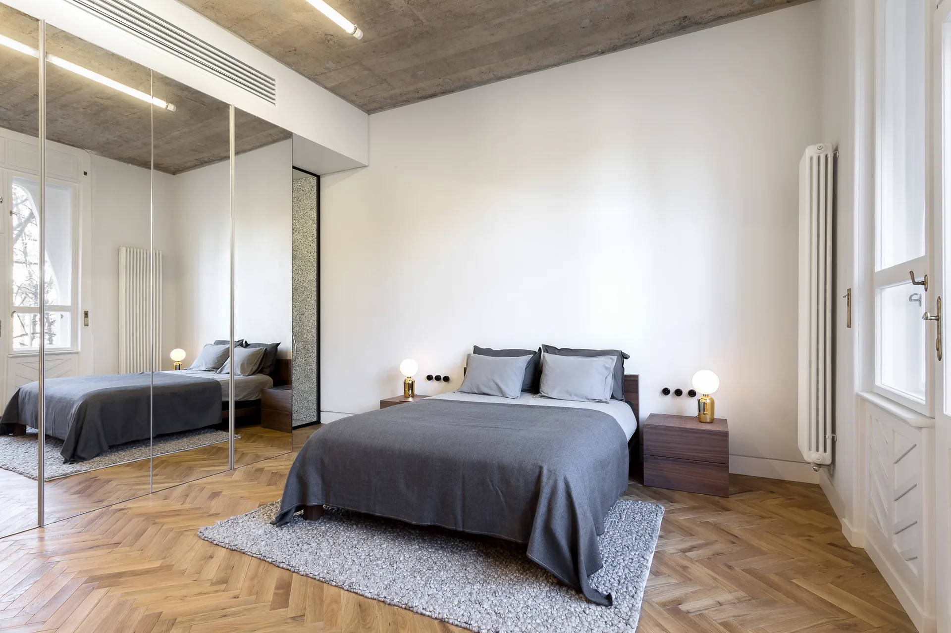 A minimalist bedroom features a bed with grey linens, wood nightstands, herringbone hardwood floors, a mirrored wardrobe, and an exposed concrete ceiling.