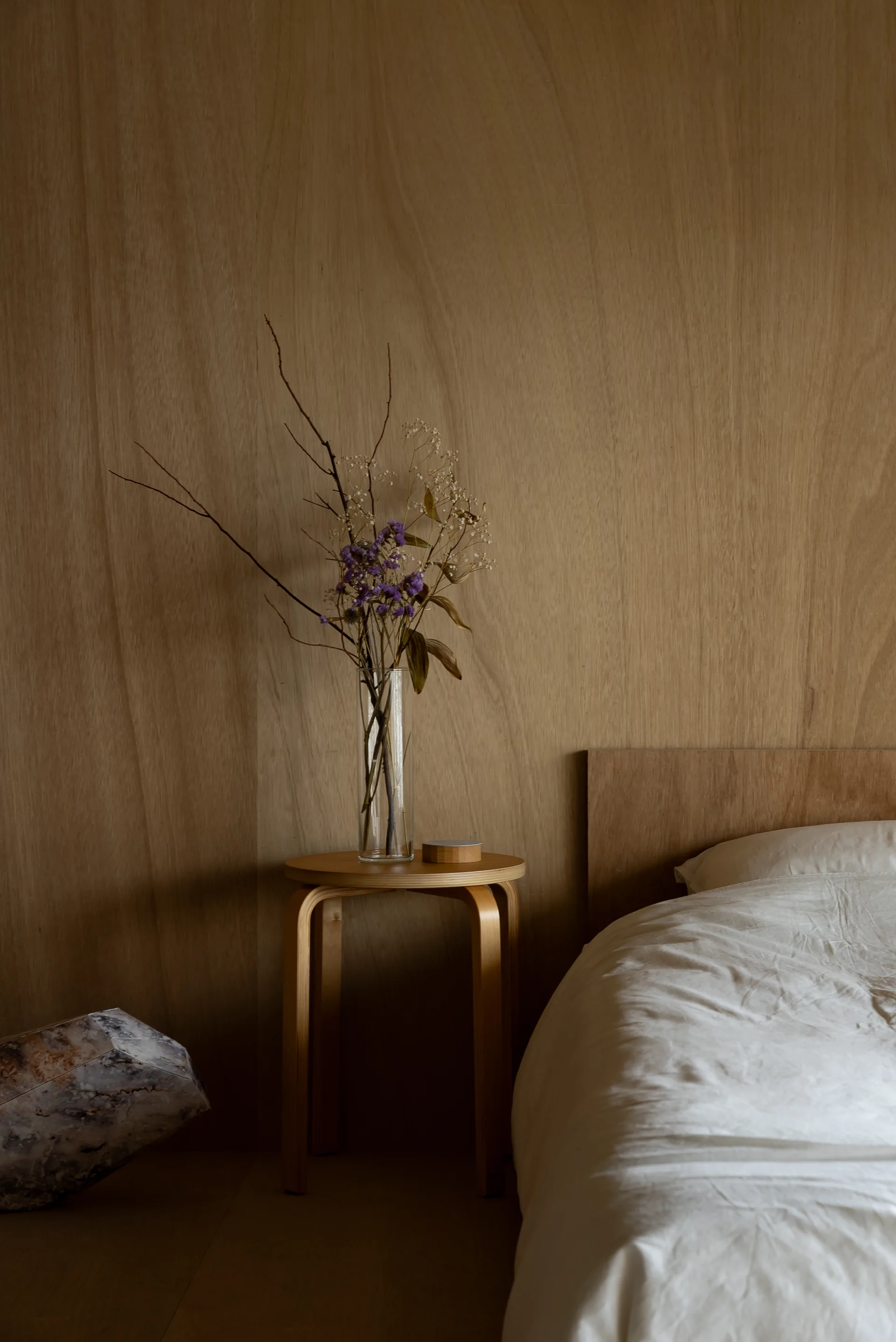 A minimalist bedroom features a warm wood paneled wall, a white bed, and a circular wooden side table holding a tall vase with dried purple flowers.