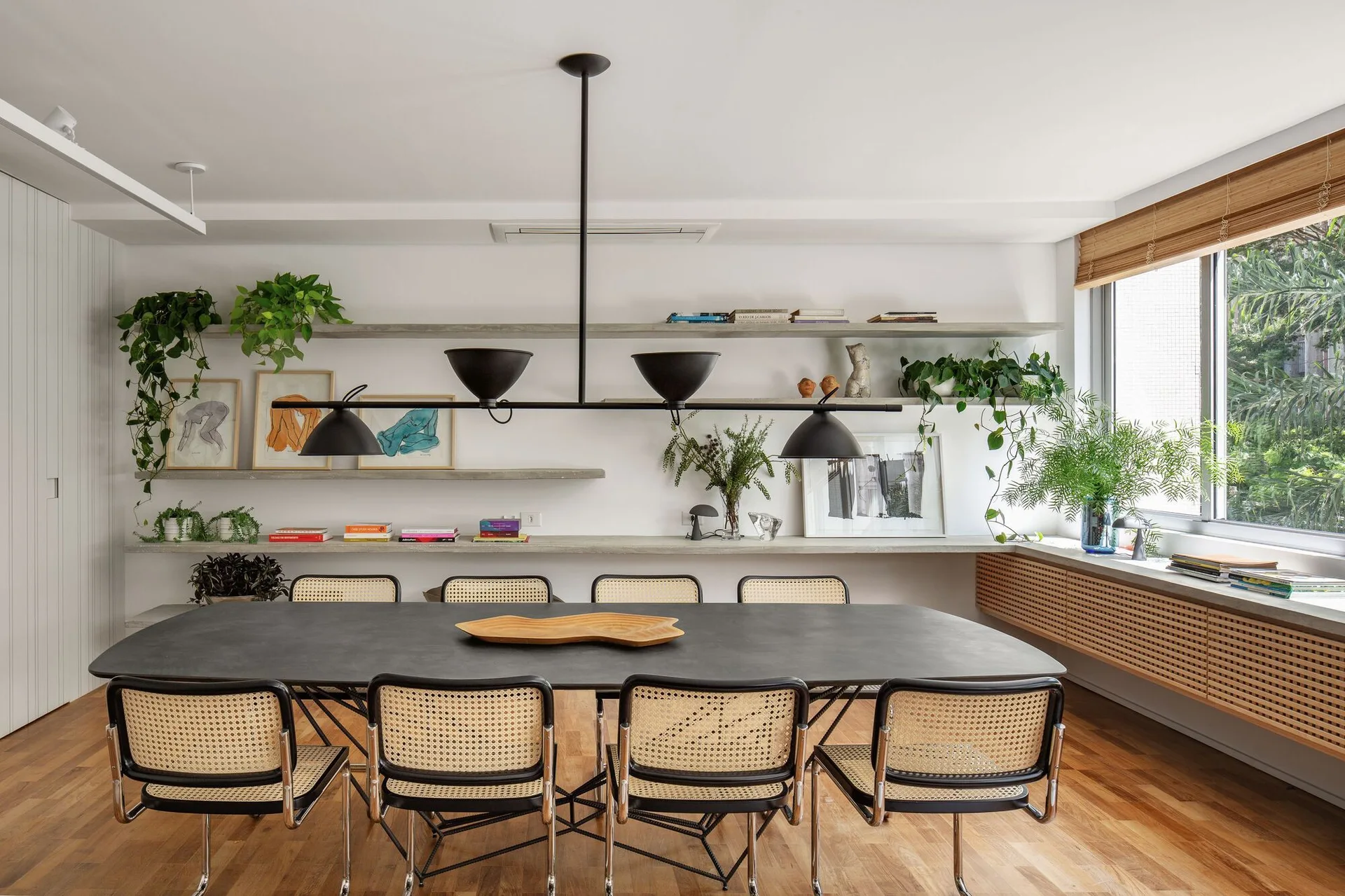A modern dining room features a dark oval table surrounded by cane-back chairs, concrete floating shelves, and a slatted wood console.