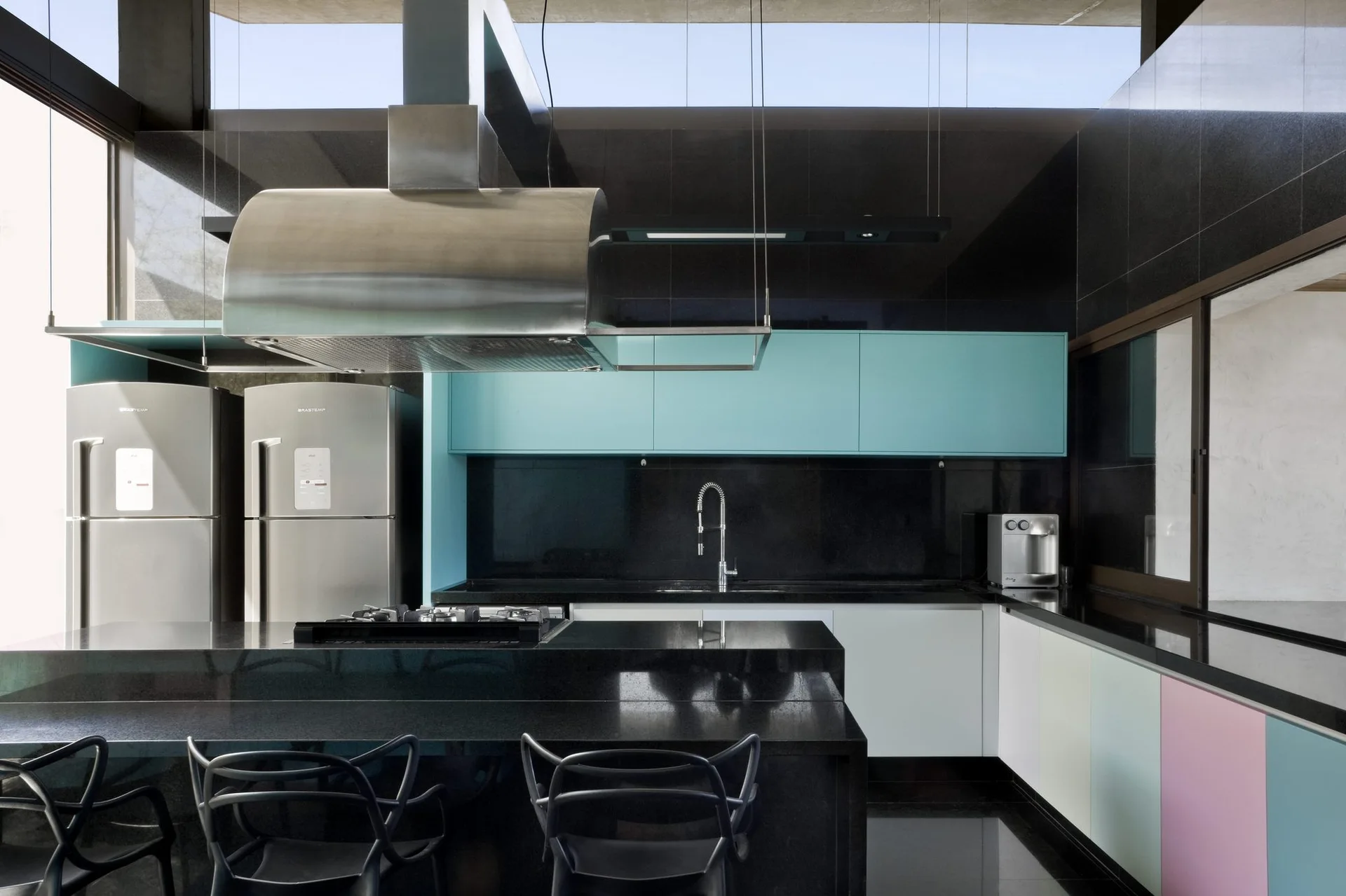 A modern kitchen features a black stone island, teal upper cabinets, pastel lower cabinetry, and two stainless steel refrigerators.