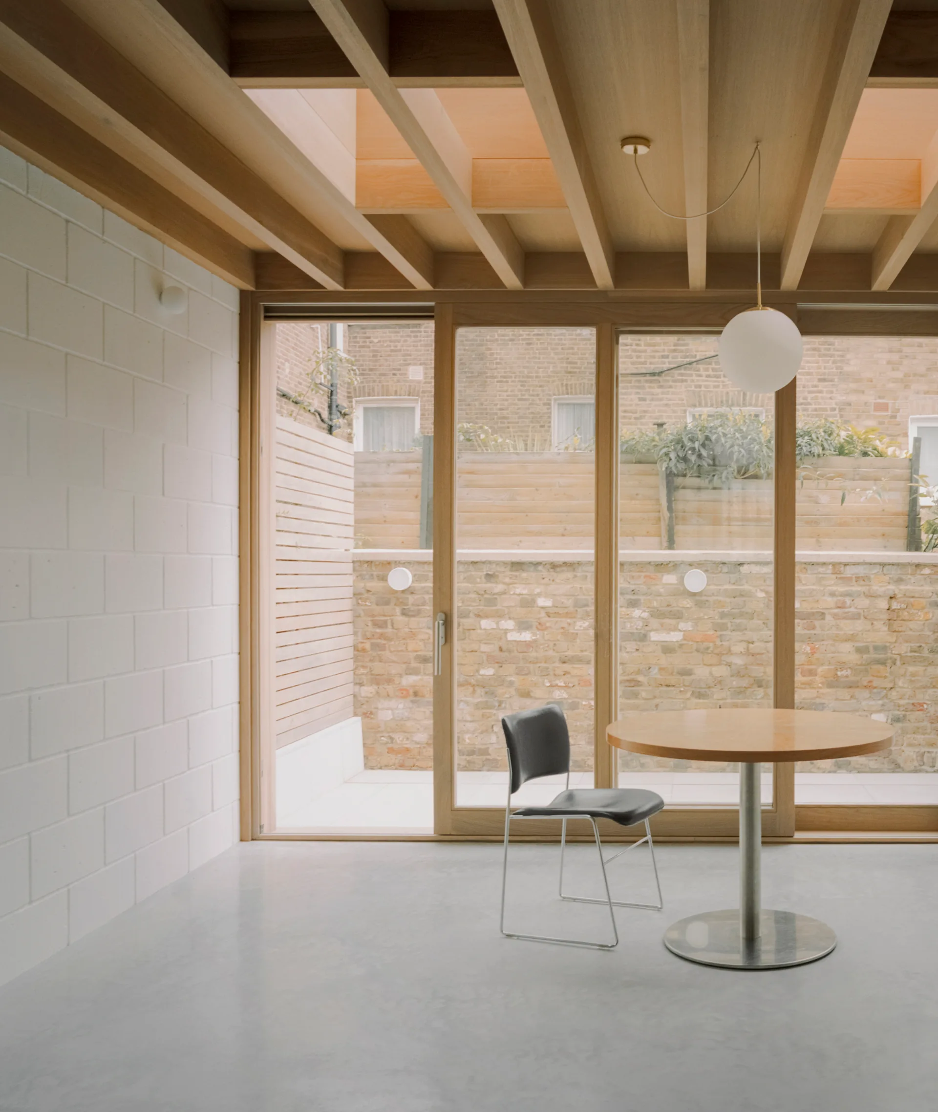 A minimalist dining space features a round wooden table, a single black chair, a polished concrete floor, and an exposed timber beam ceiling.