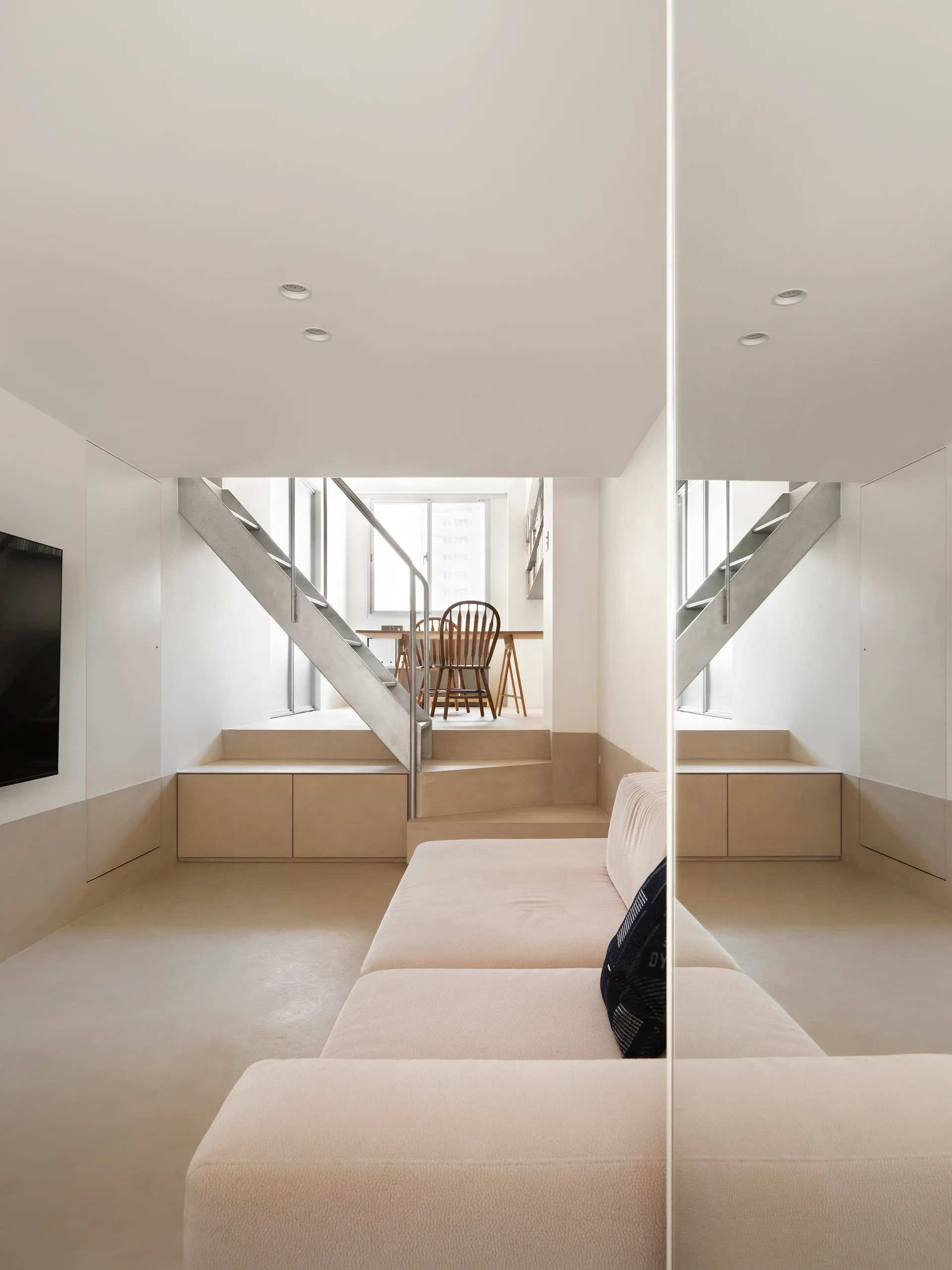 A minimalist split-level living room features a beige sofa, metal stairs leading to a raised dining area, and a large mirrored wall on the right.