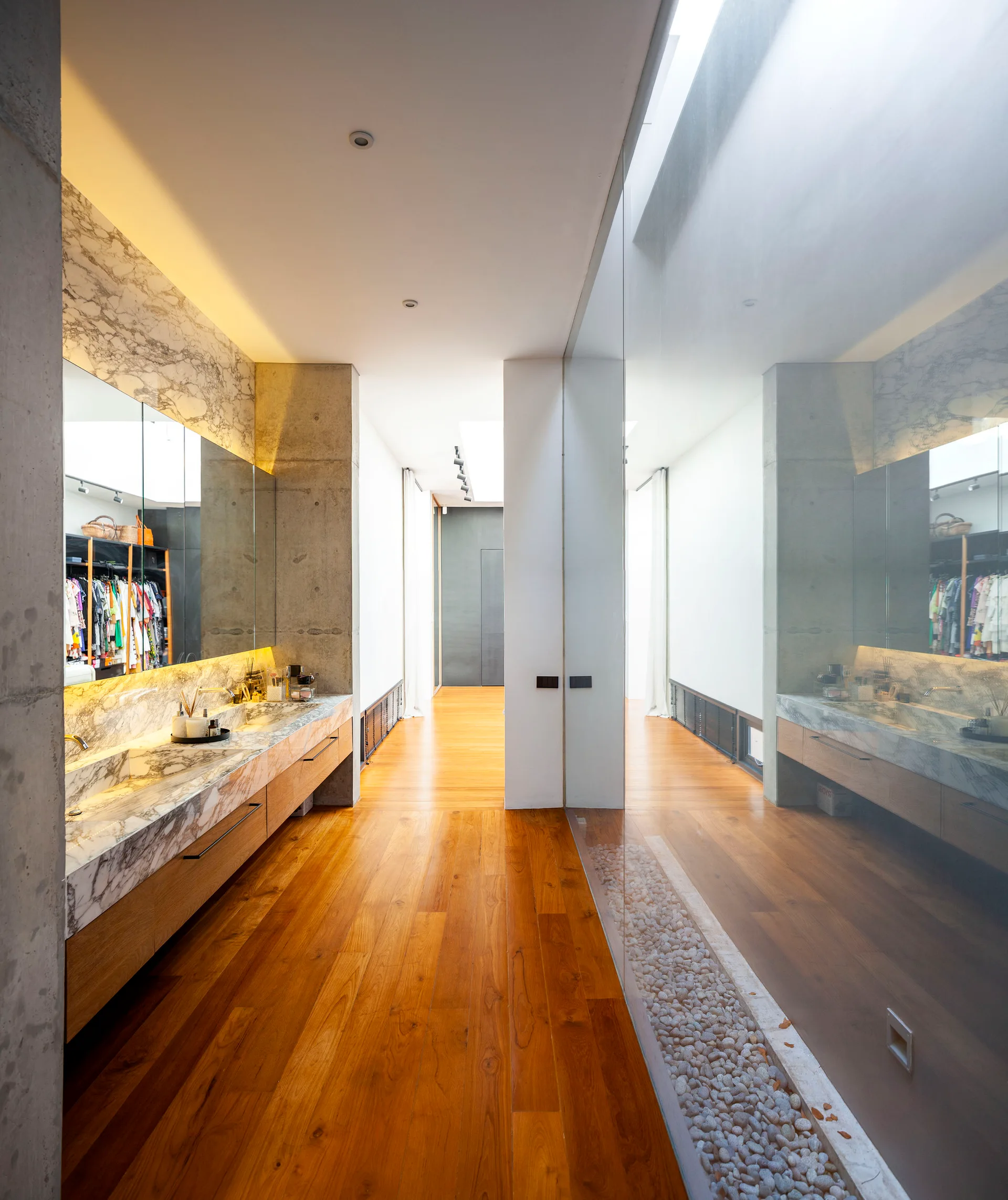 A modern minimalist bathroom featuring a floating marble vanity, warm wood plank flooring, and a linear stone pebble trench alongside a glass wall.