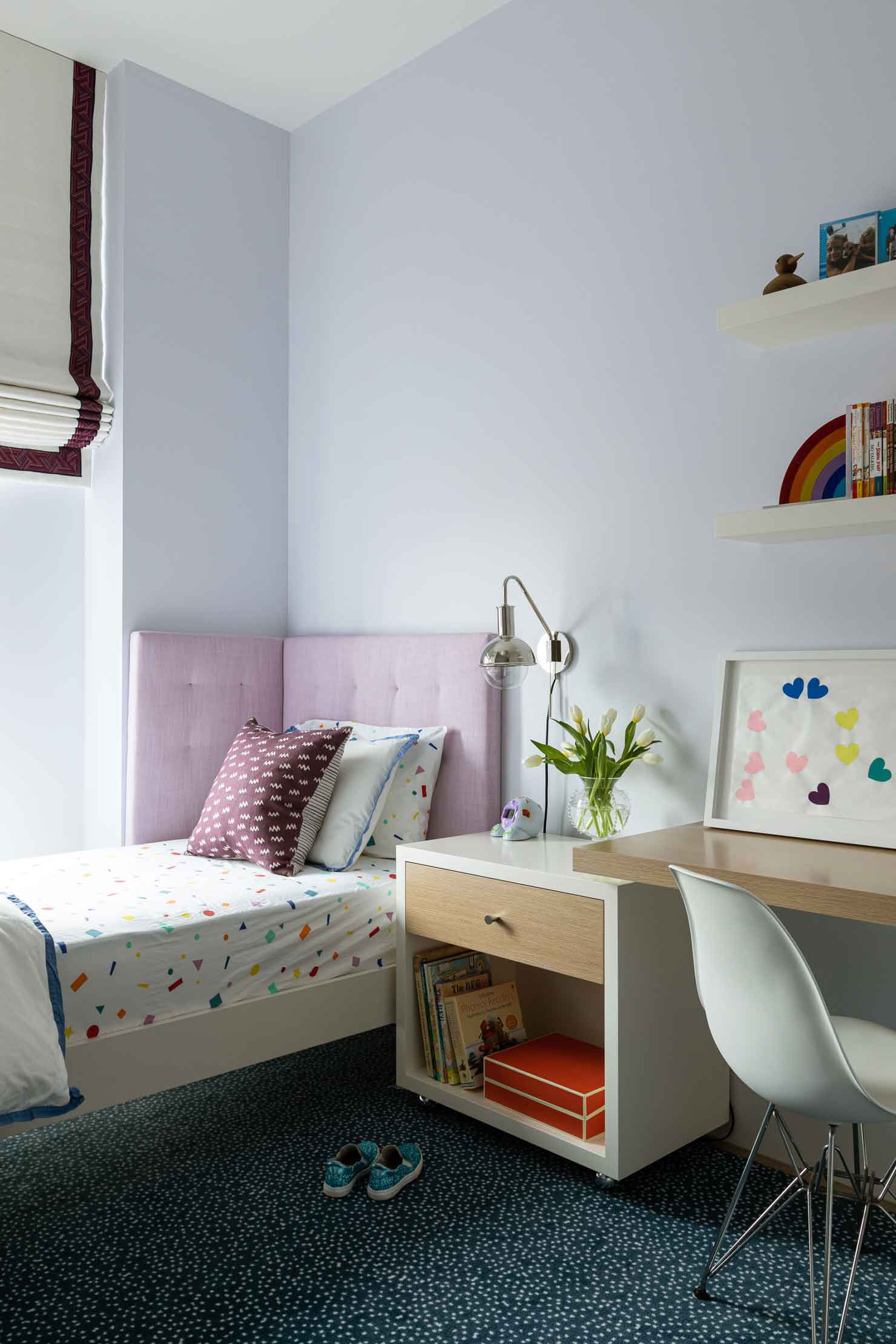 A kids bedroom features a single bed with a lavender corner headboard, a white rolling nightstand, a wood desk with a white chair, and floating shelves.