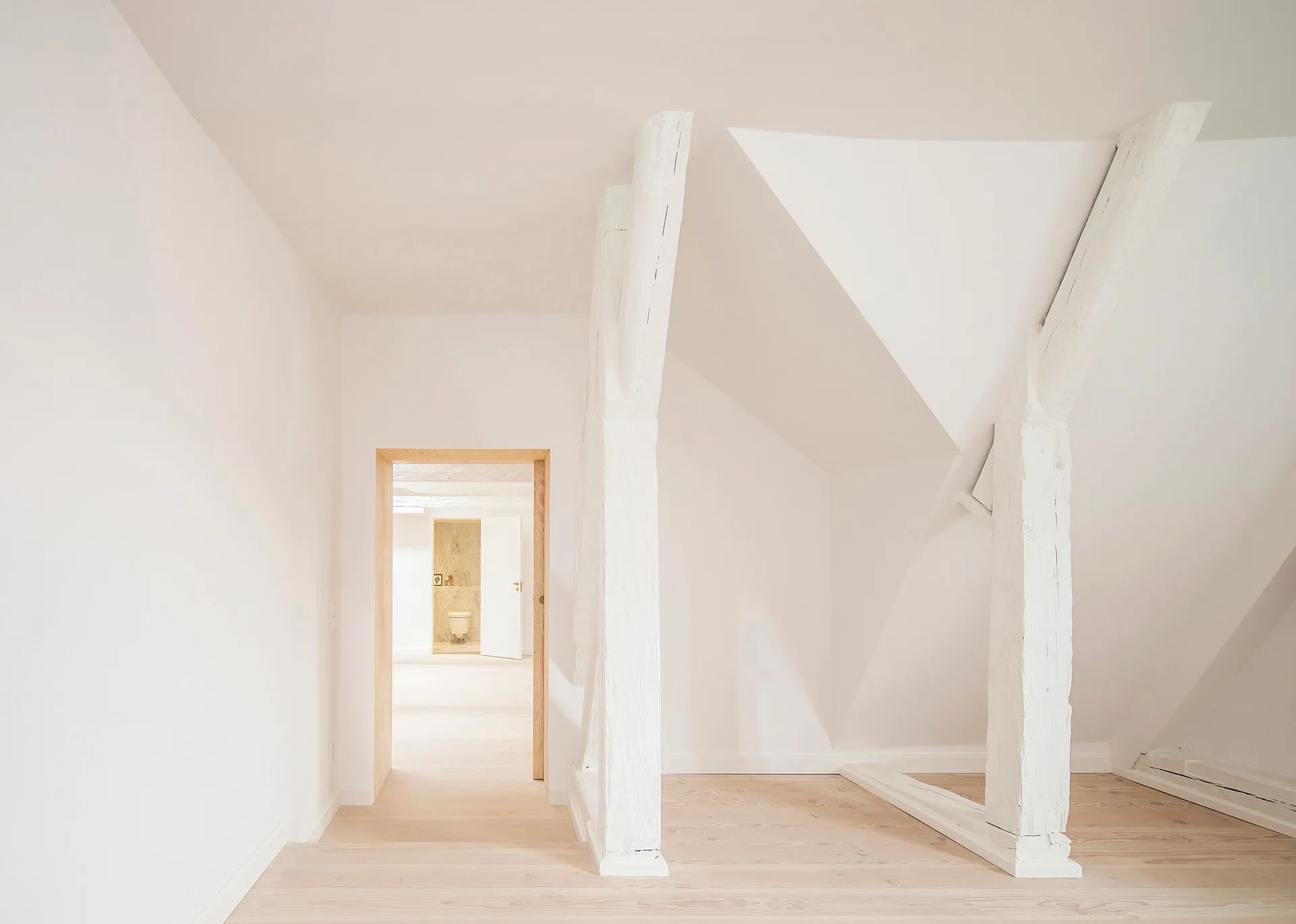 A minimalist white attic hallway features painted timber structural posts, light wood floors, and an open doorway leading to another room.
