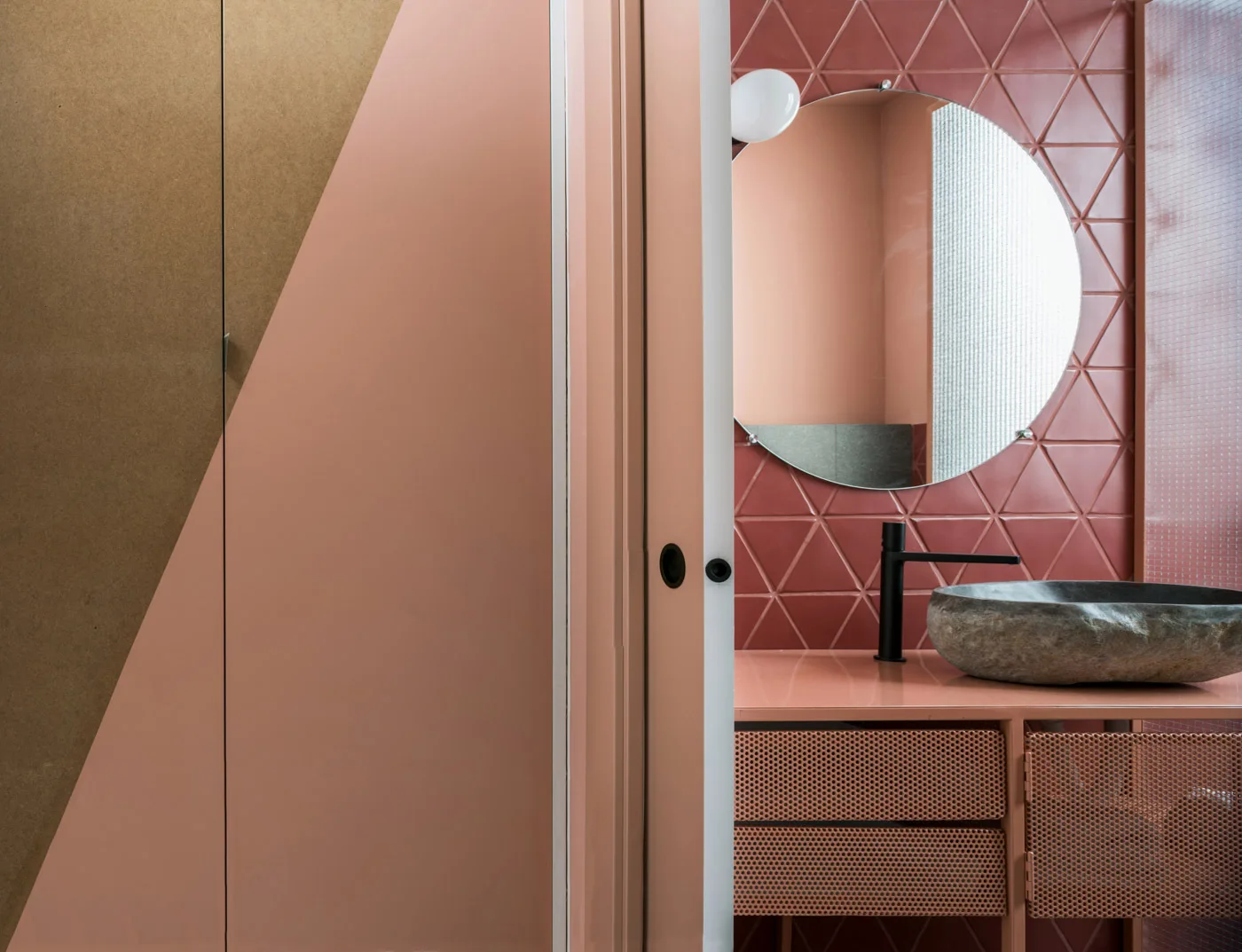 A modern bathroom vanity with a rough stone vessel sink, matte black faucet, and a round mirror against pink triangular wall tiles.