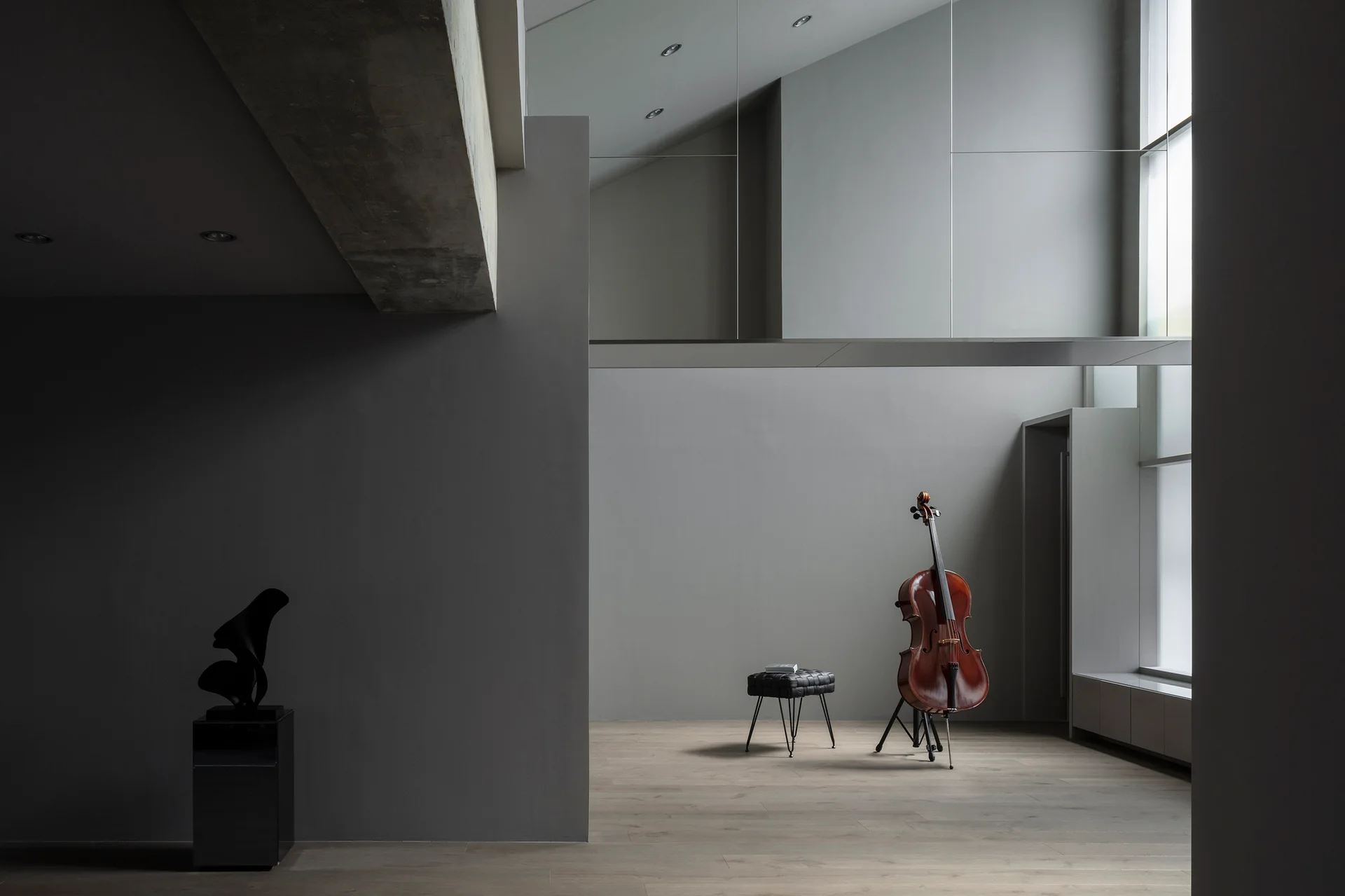 A minimalist gray room features a cello on a stand and a black tufted stool on wood flooring near a large window.