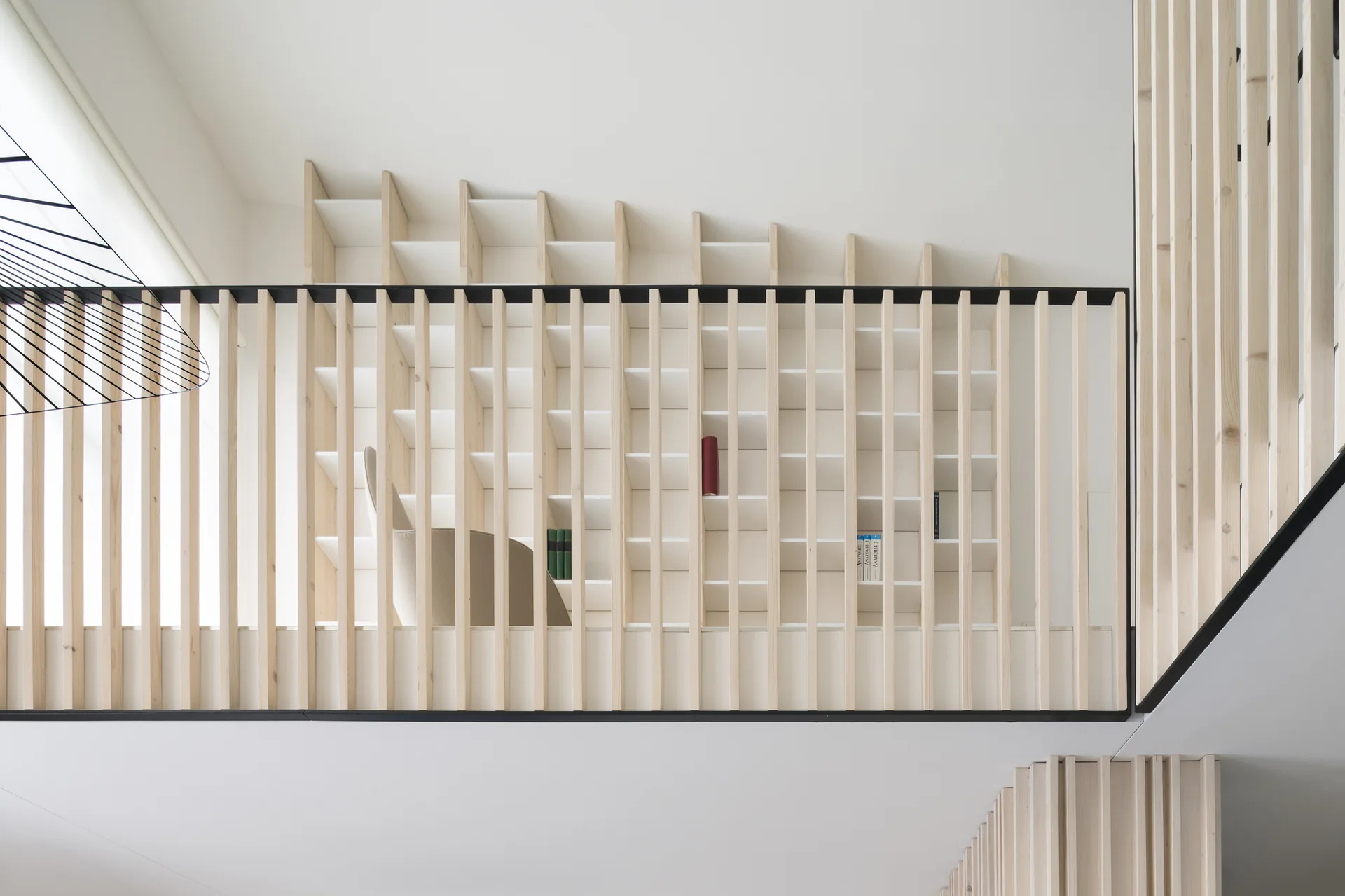 A low-angle view of a mezzanine landing featuring a vertical light wood slatted railing in front of a built-in grid bookshelf and white walls.