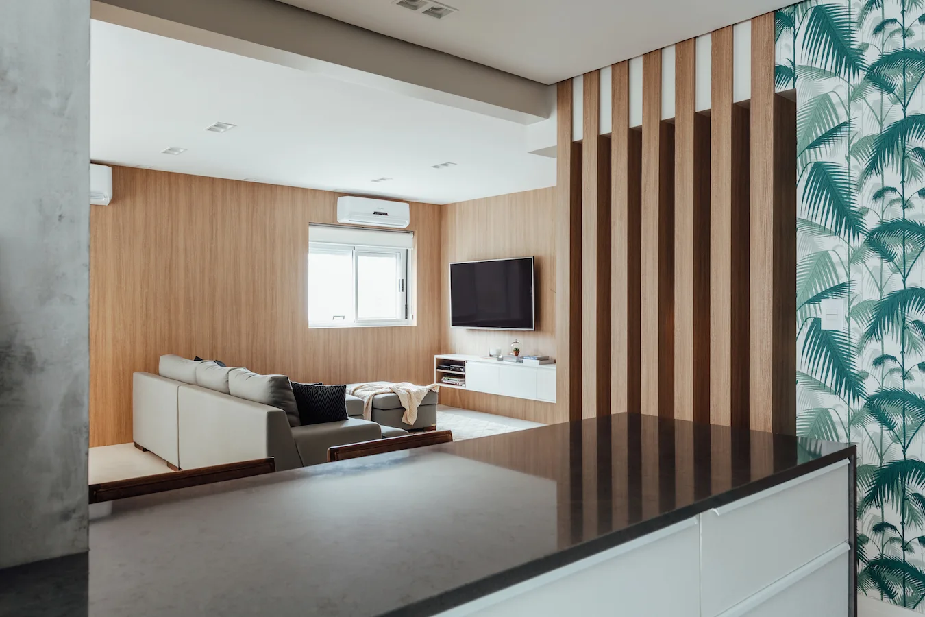 A modern living room featuring wood-paneled walls, a gray sectional sofa, a white floating media console, a slatted wood divider, and green palm wallpaper.