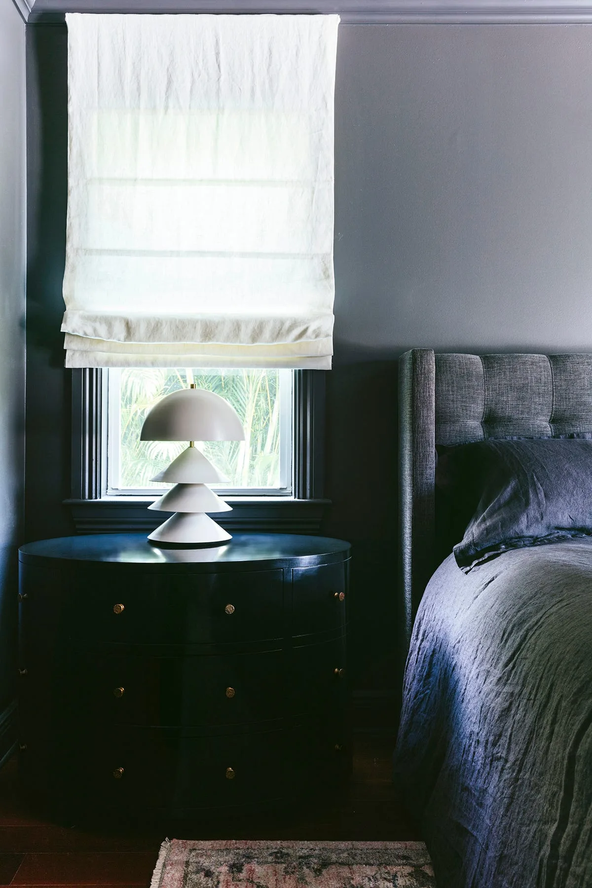 A dark charcoal bedroom corner featuring a round black three-drawer nightstand, a white tiered table lamp, and a white fabric roman shade.
