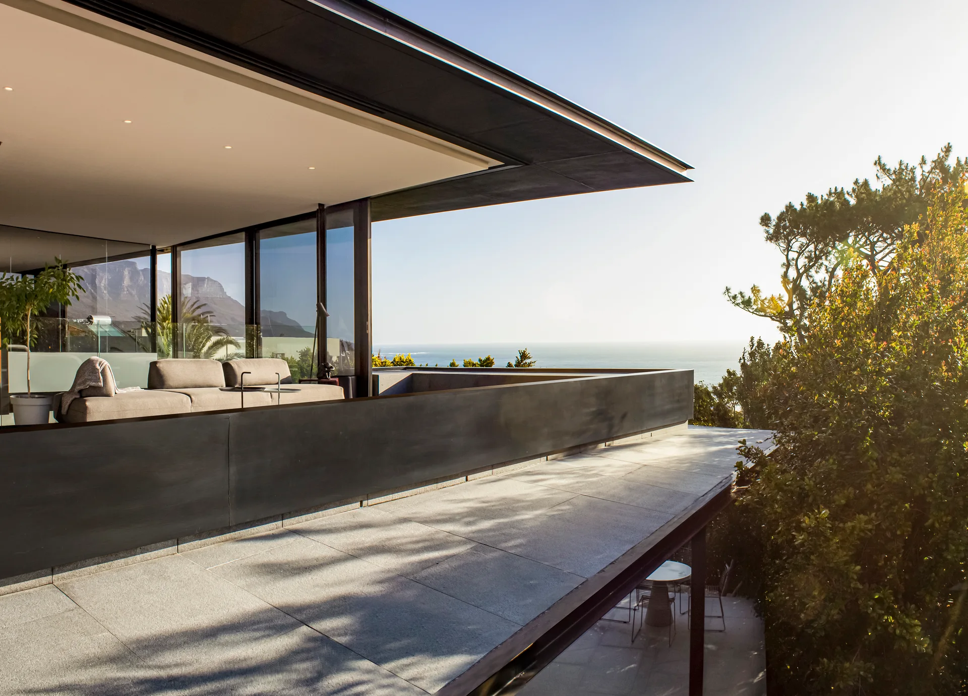A modern terrace features a modular sofa under a cantilevered roof next to a stone-tiled floor and a dark metal railing overlooking the sea.