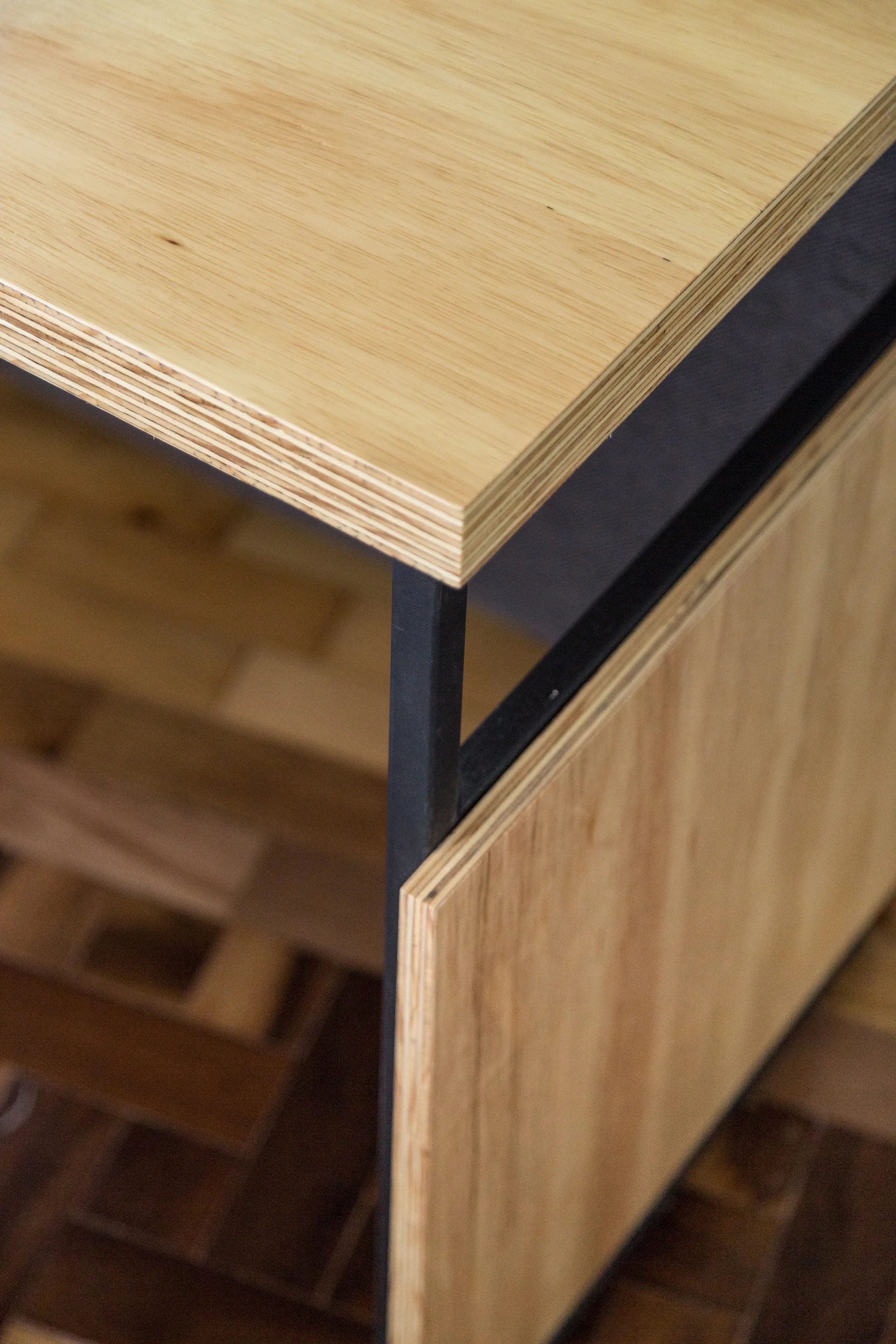 A close-up of a light wood table corner featuring exposed plywood edges and a matte black metal frame over a parquet floor.