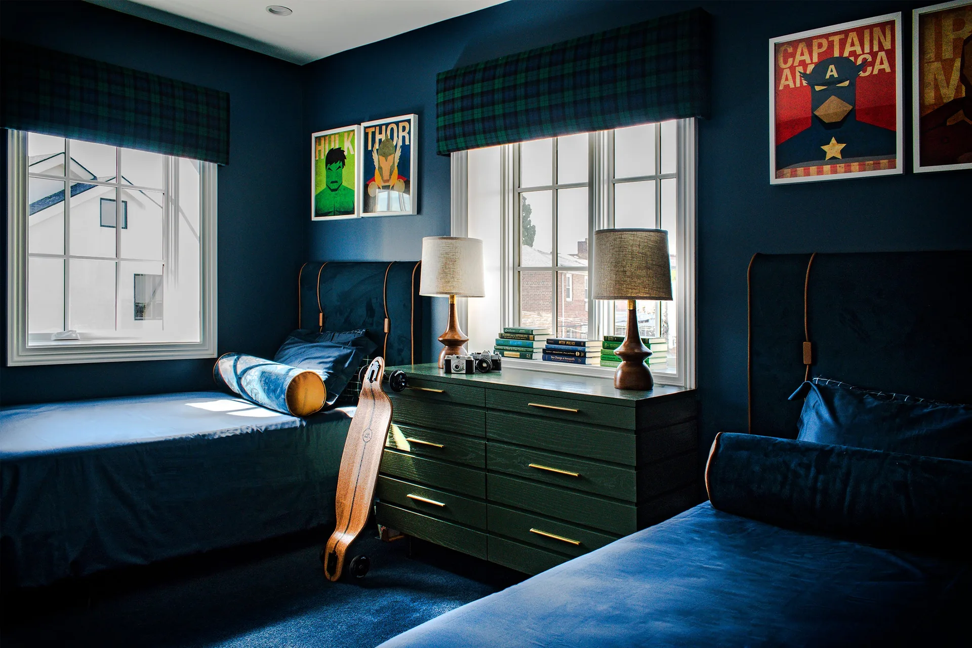 A shared children's bedroom features navy blue walls, matching upholstered twin beds, and a forest green dresser under a window with plaid valances.