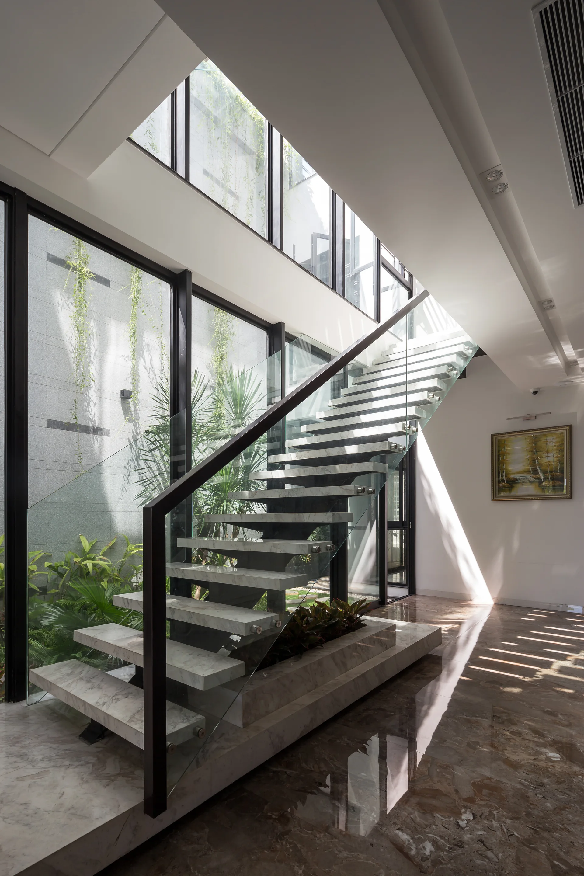 A modern floating staircase with marble treads and glass railing sits beside a glass-enclosed internal courtyard filled with green plants.