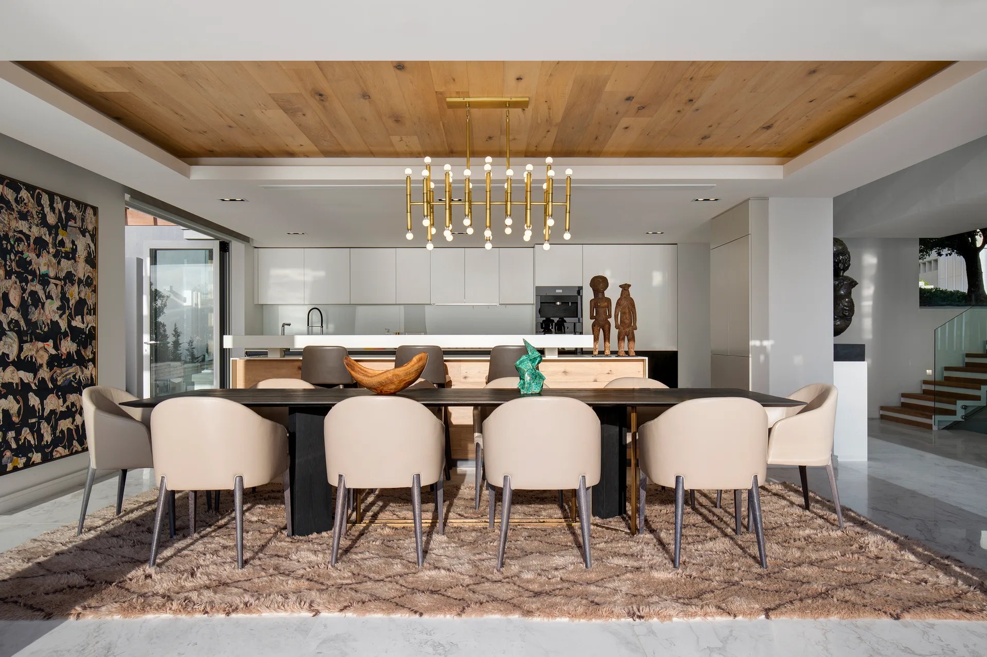 A contemporary dining room with a long dark wood table, beige leather chairs, a textured shag rug, and a recessed wood plank ceiling.