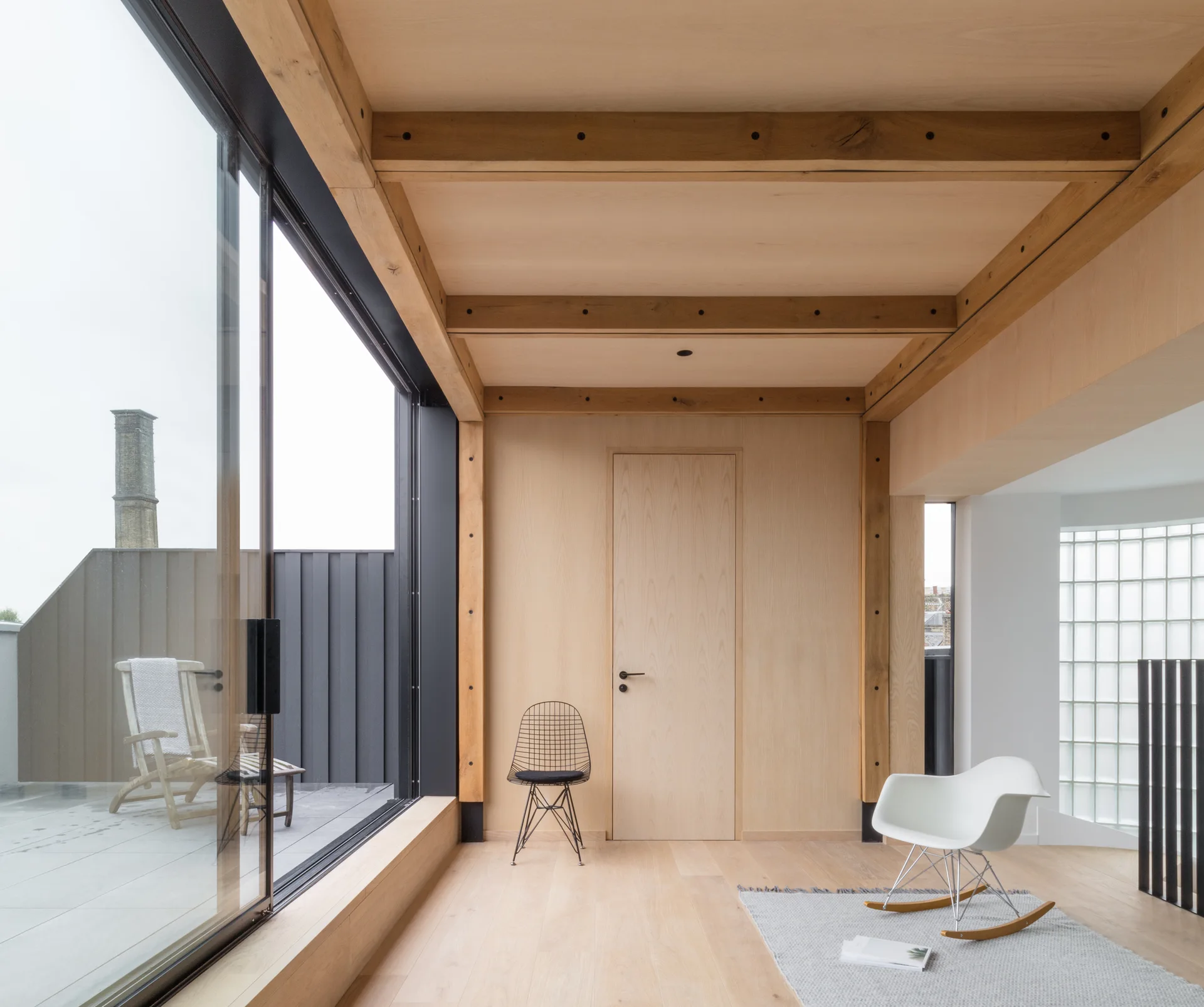 A minimalist room featuring light wood paneled walls and ceiling beams, a sliding glass door leading to a terrace, a grey rug, and two modern chairs.
