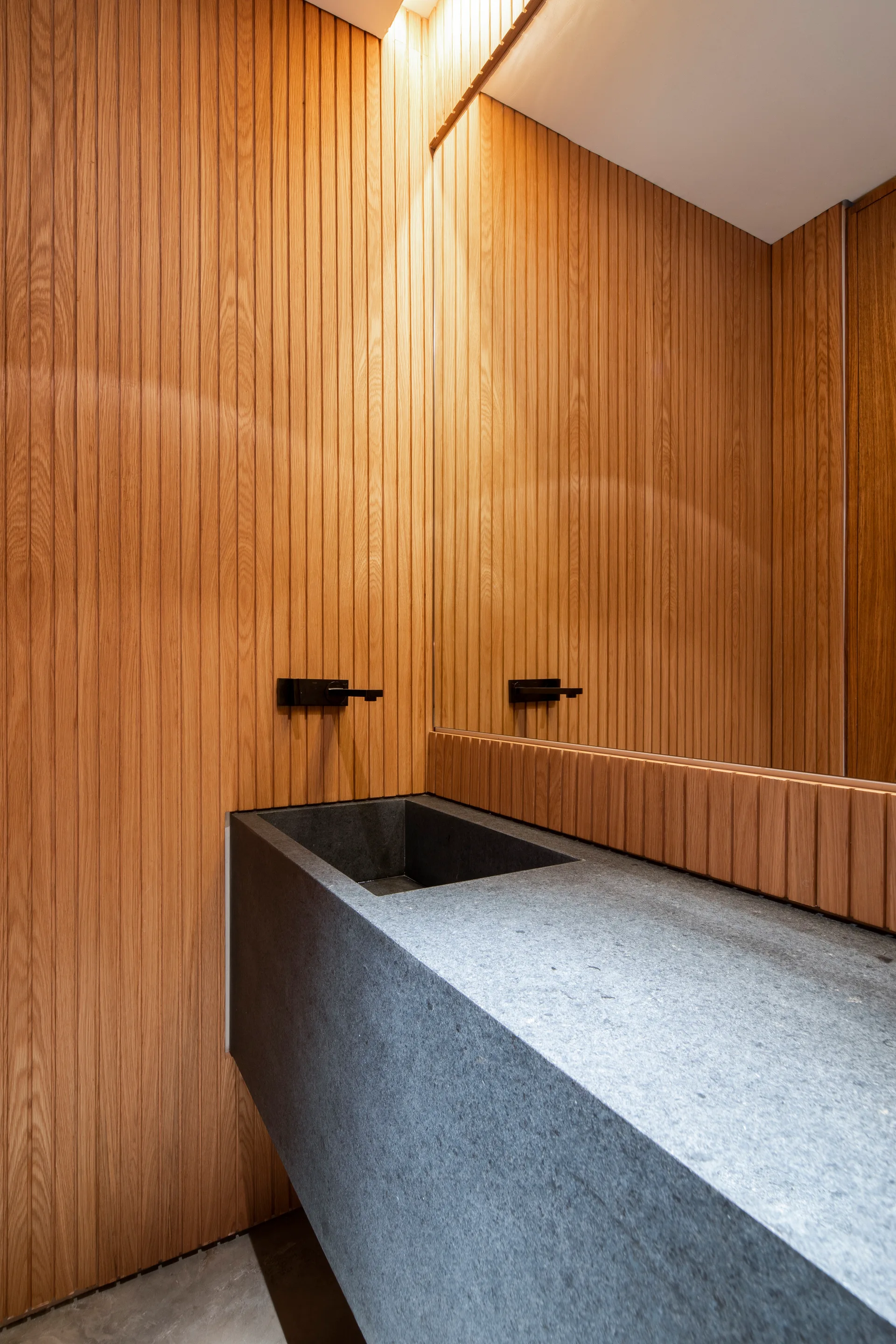 A modern powder room features a large grey stone floating sink, vertical wood slat wall paneling, a black wall-mounted faucet, and a large mirror.