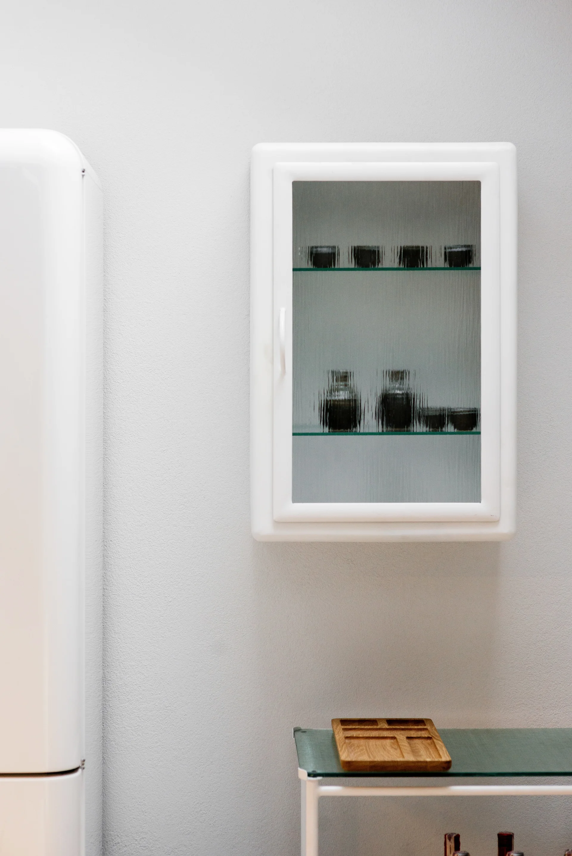 A white wall-mounted cabinet with a fluted glass door hangs above a glass console table next to a retro refrigerator.