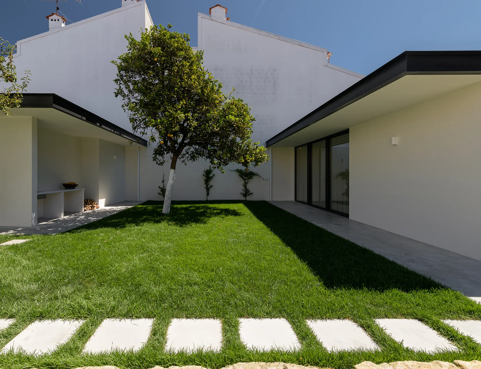 A minimalist courtyard features a green lawn, a stone stepper path, a single central tree, and white exterior walls.