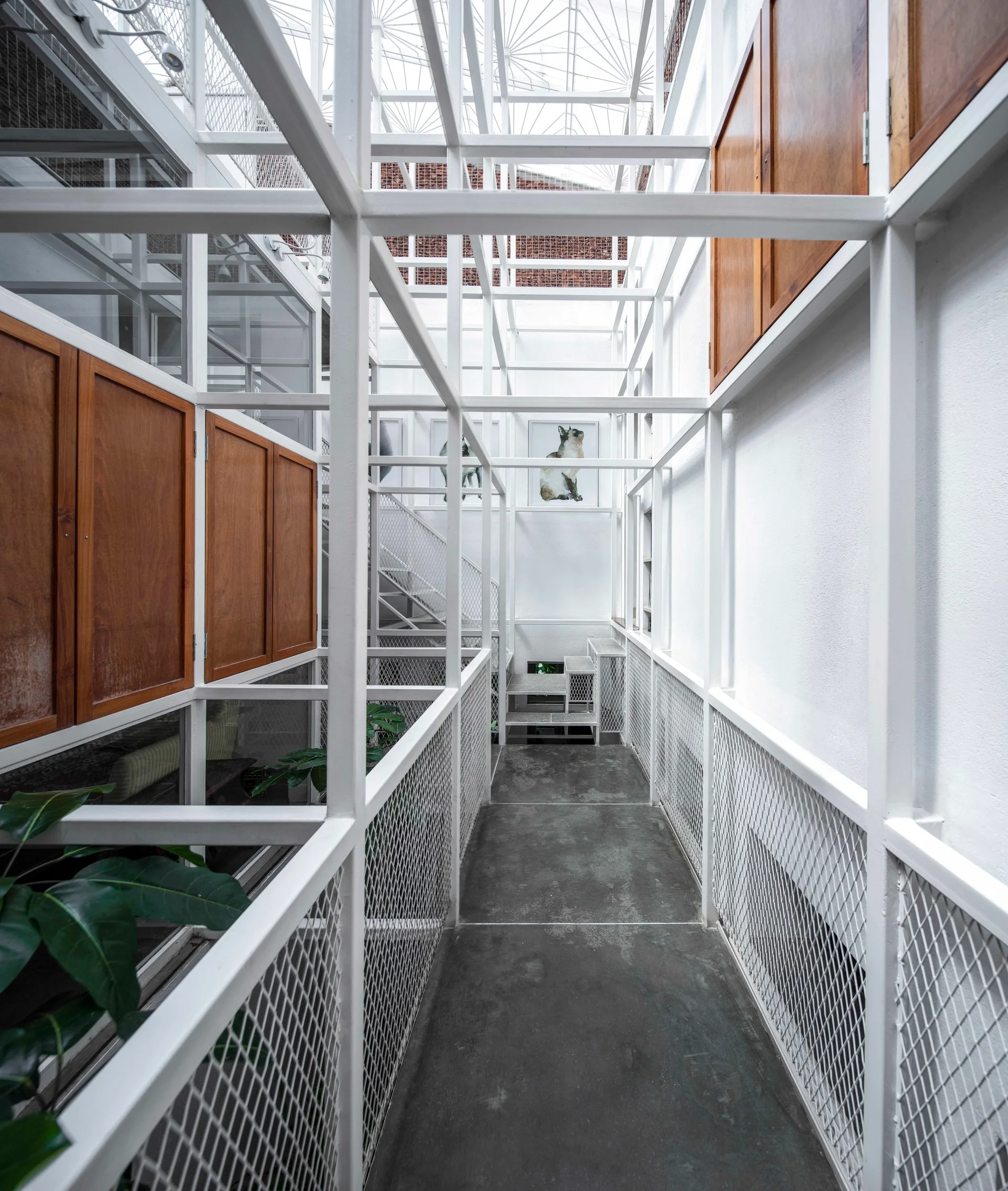 A narrow concrete walkway is surrounded by a white metal grid structural frame with built-in wood cabinets and mesh railings under a skylight.