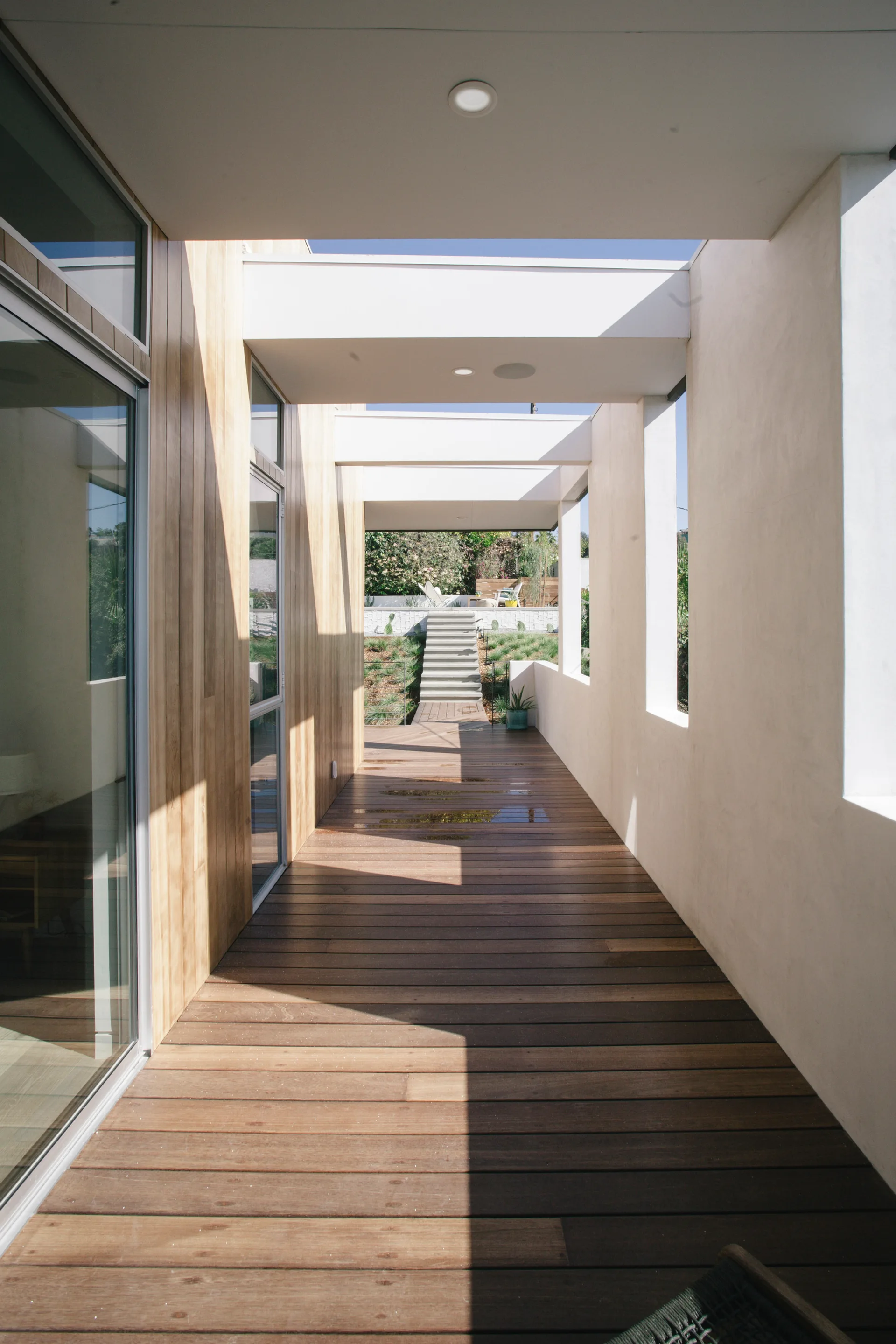 A modern outdoor hallway featuring a brown wood plank deck, a vertical wood siding wall, and white stucco columns leading to concrete garden steps.
