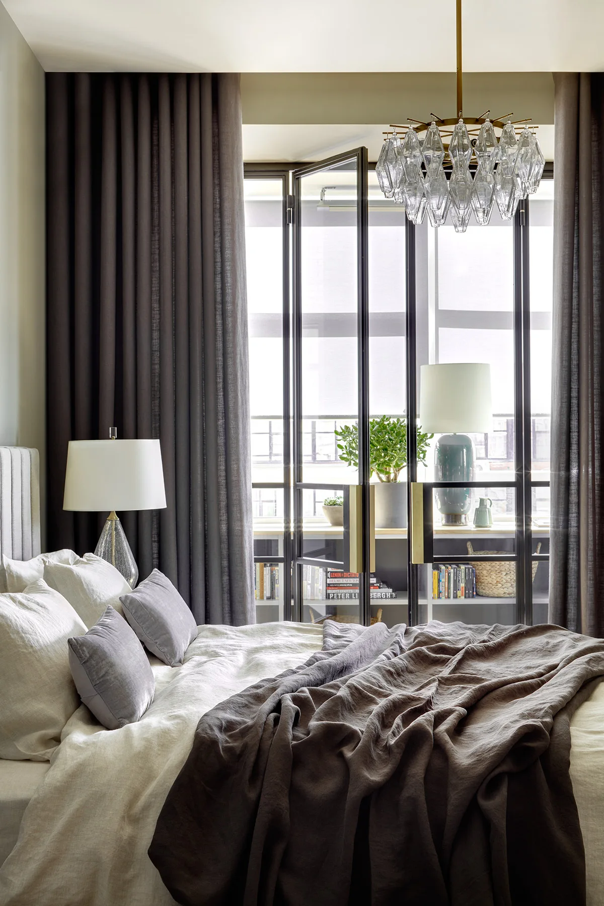 A bedroom featuring neutral layered bedding, a crystal chandelier, and dark drapes next to glass-paned folding doors that divide the space.