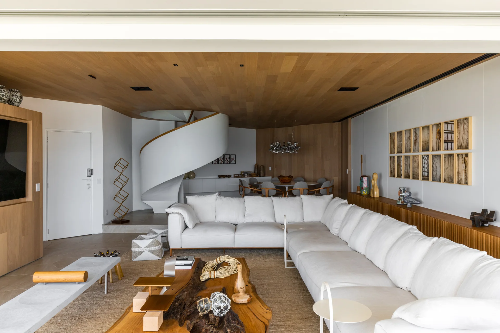 A spacious contemporary living room features a cream sectional sofa, a live-edge wooden coffee table, a wood plank ceiling, and a white spiral staircase.