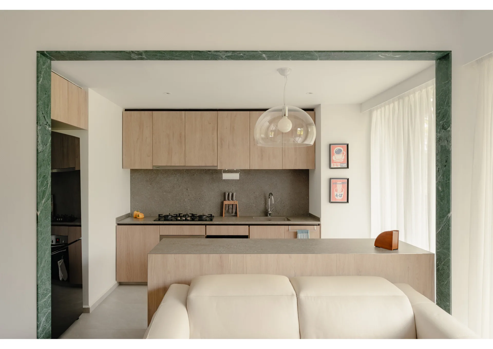 A minimalist kitchen with light wood cabinets and stone countertops, viewed through a thick green marble frame with a cream sofa in the foreground.